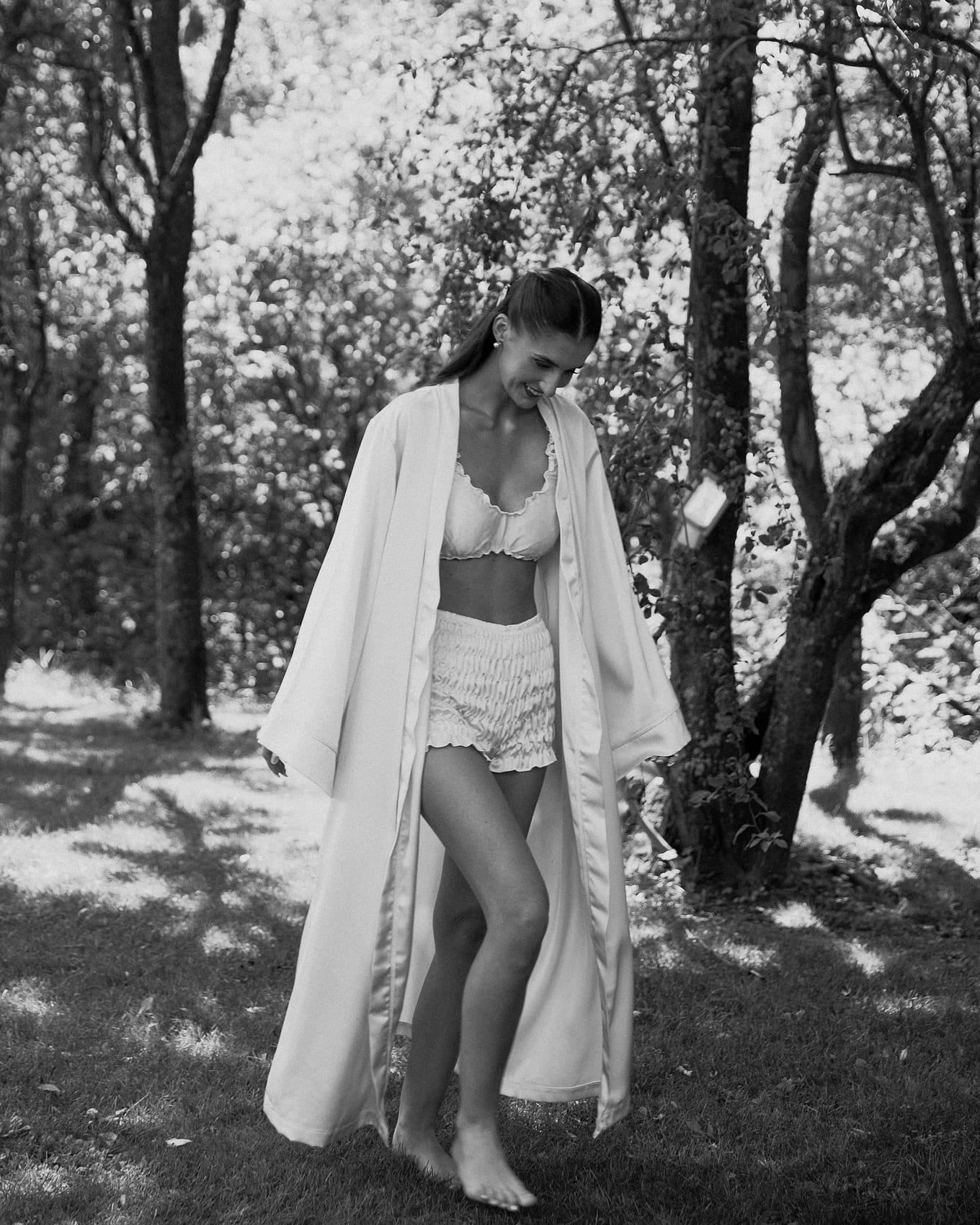 Smiling, a woman in a long open robe and ruffled shorts walks barefoot across shaded grass