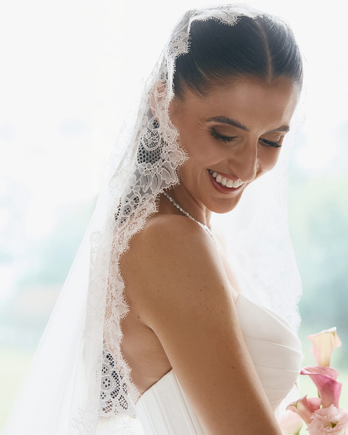 Smiling bride with a lace-edged veil looks down toward pink calla lilies by a bright window
