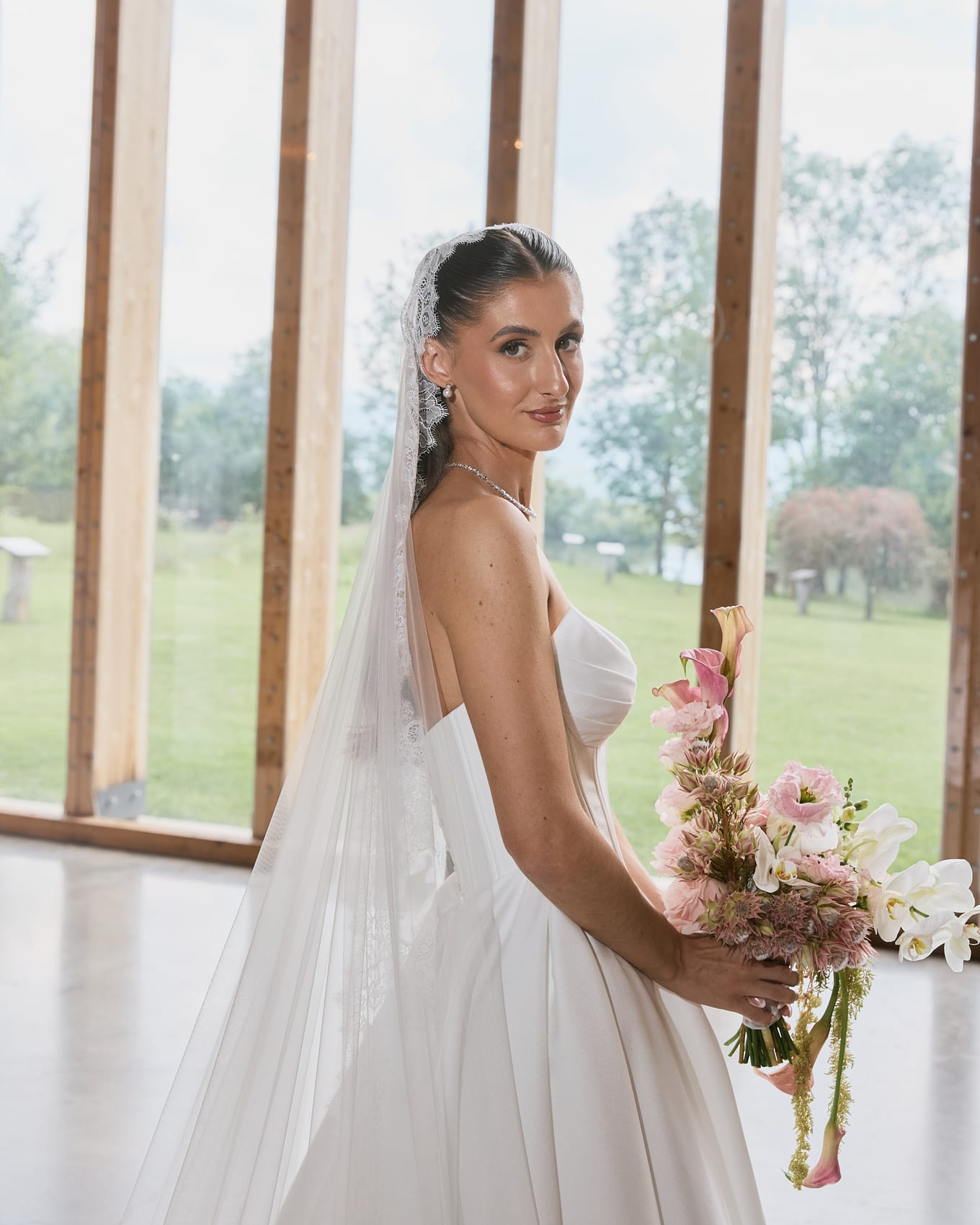 Bride holding a pink and white bouquet looks over her shoulder beside tall windows facing a lawn