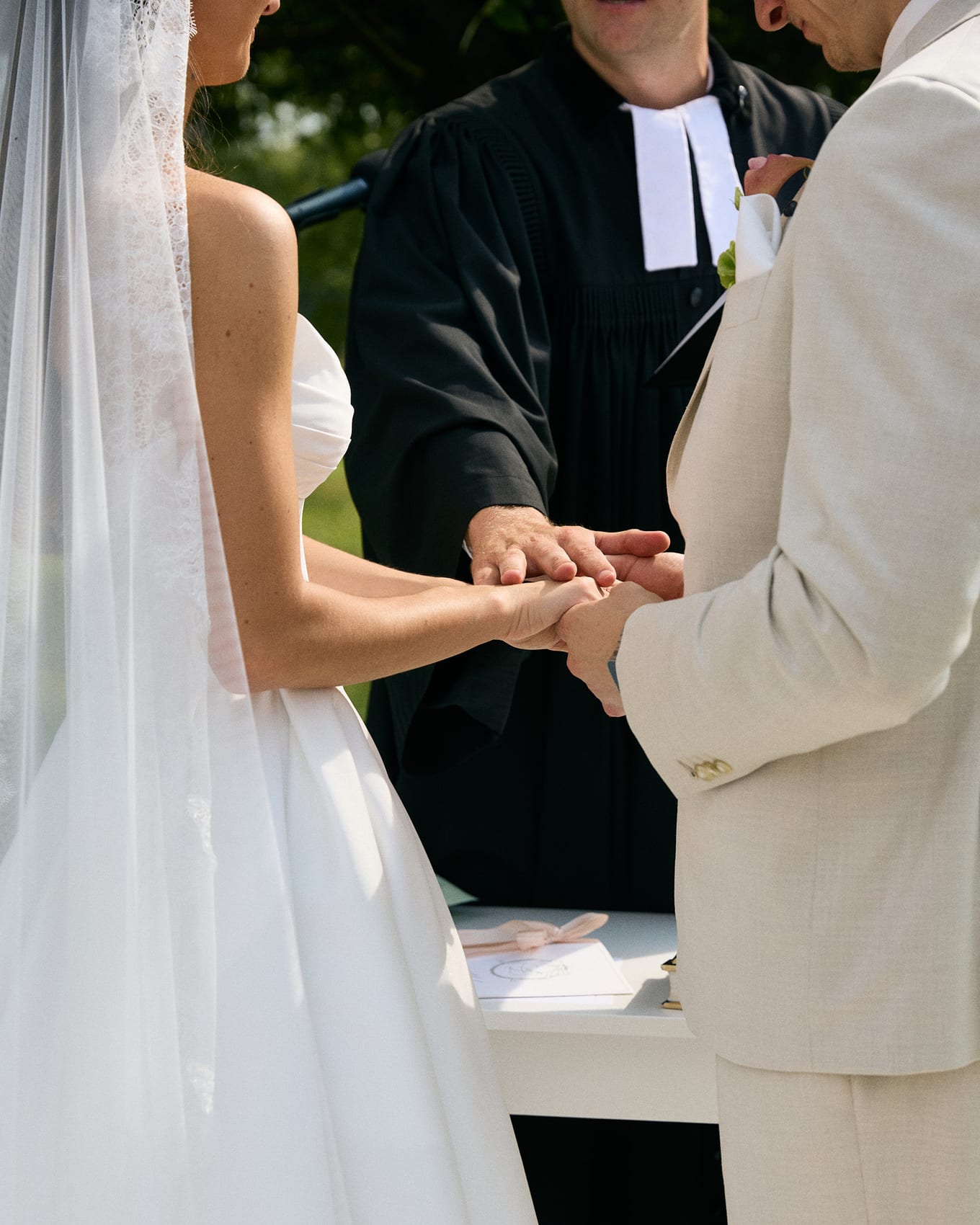 Close-up of an officiant in a black robe resting a hand on the joined hands of the bride and groom
