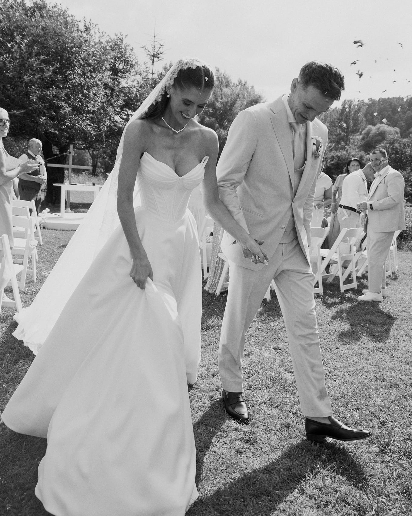 Bride lifting her gown and smiling as she walks hand in hand with the groom past white chairs