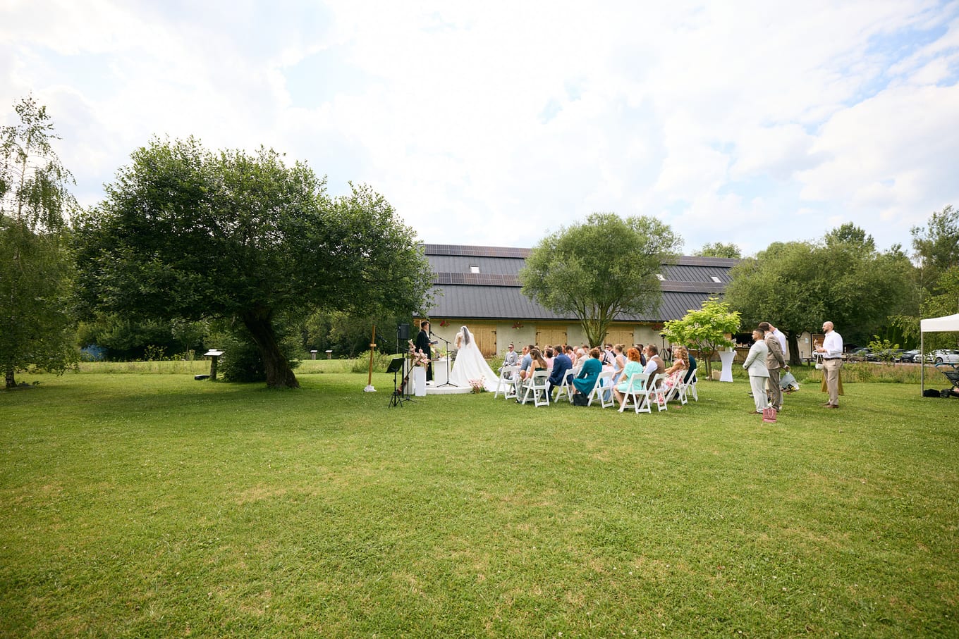 Outdoor ceremony on a wide lawn, guests seated in white chairs before a long dark-roofed building