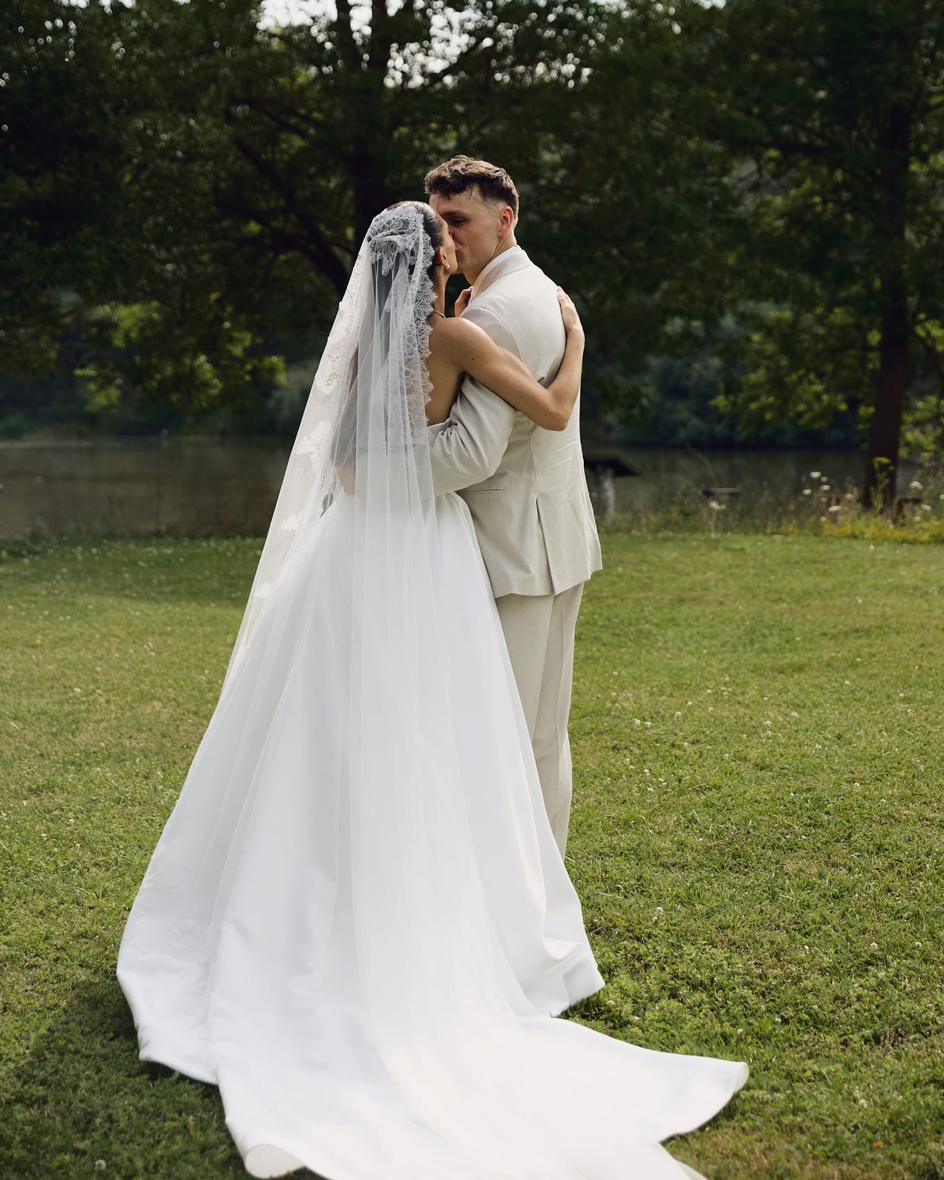 Bride in a lace-edged veil embraces the groom in a kiss on a lawn beside a pond