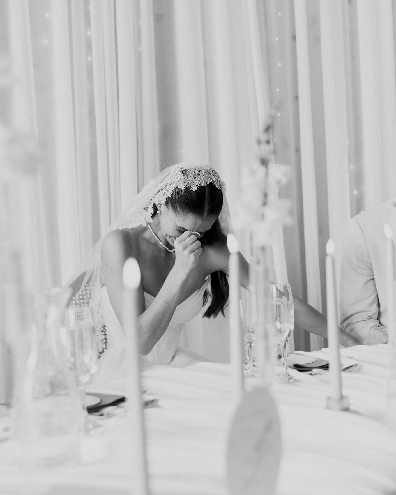 Seated at a candlelit table, the bride lowers her head and wipes her eyes with one hand