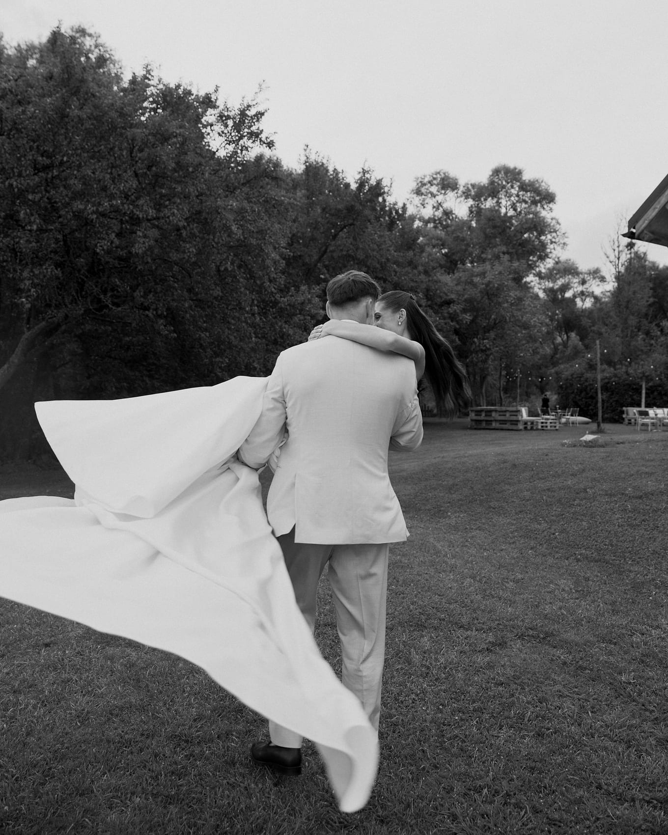 Groom in a pale suit carries the bride across a lawn as her long train billows behind them