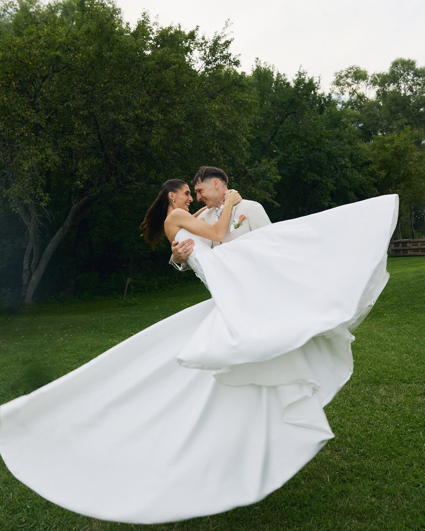 Laughing bride held in the groom's arms on green grass, her white gown flaring out in a wide arc