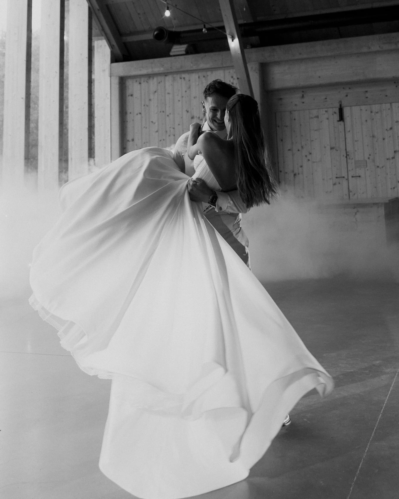 Inside a wooden hall filled with low fog, the groom swings the bride as her skirt sweeps around