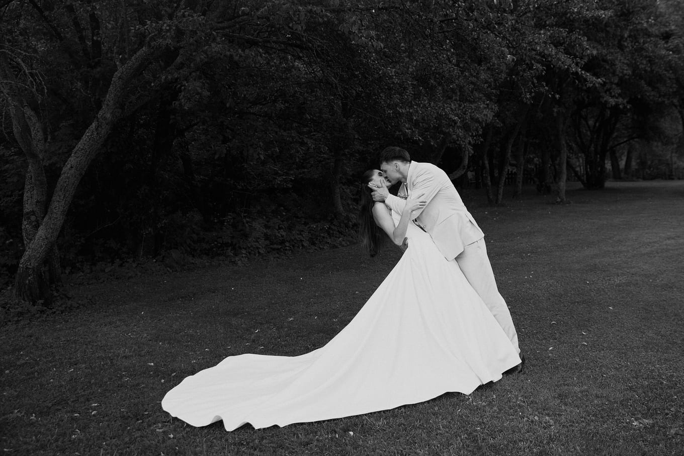 Groom dipping the bride for a kiss on a lawn, her long train spread across the grass before dark trees