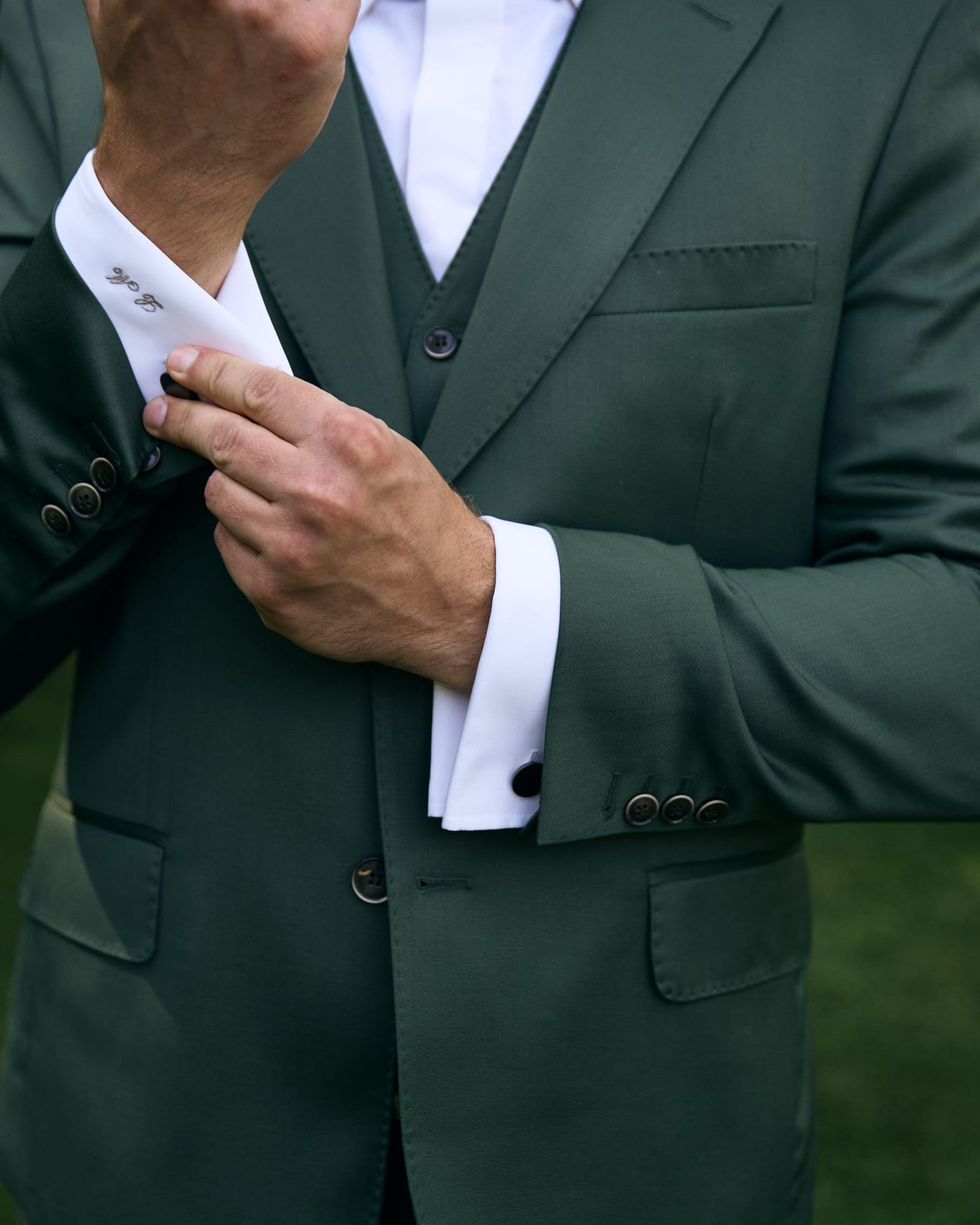 Close-up of a hand adjusting a monogrammed white shirt cuff beneath a green suit sleeve