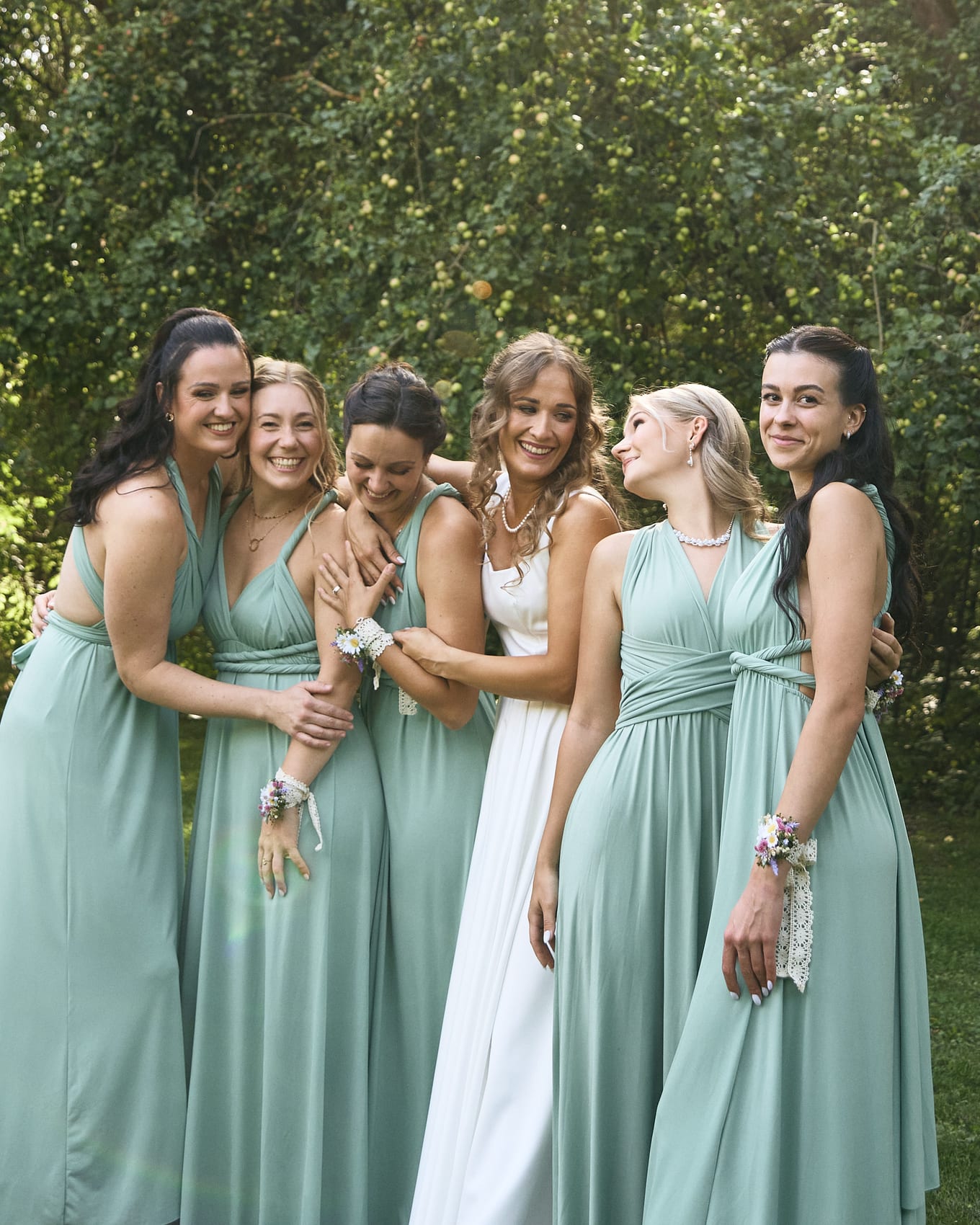 Six women in sage and white dresses hug and smile together in front of leafy green bushes