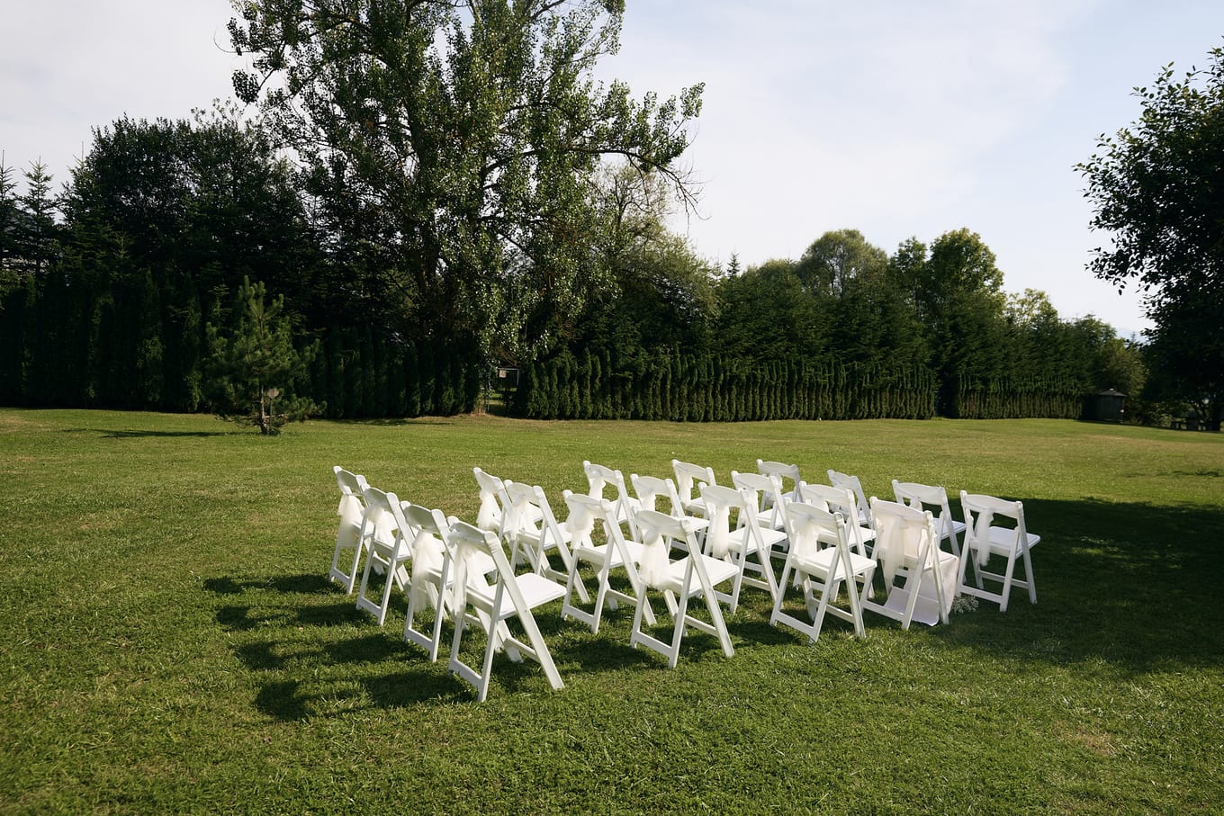 Rows of white folding chairs set out on a mowed lawn in front of tall trees and a hedge