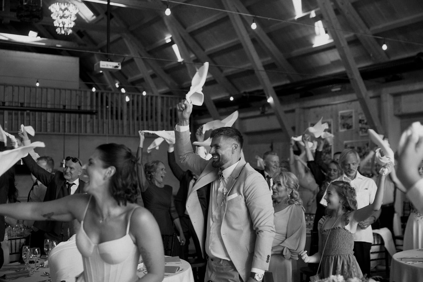 Guests wave white napkins overhead in a wooden-beamed hall, a smiling man in a light suit at centre