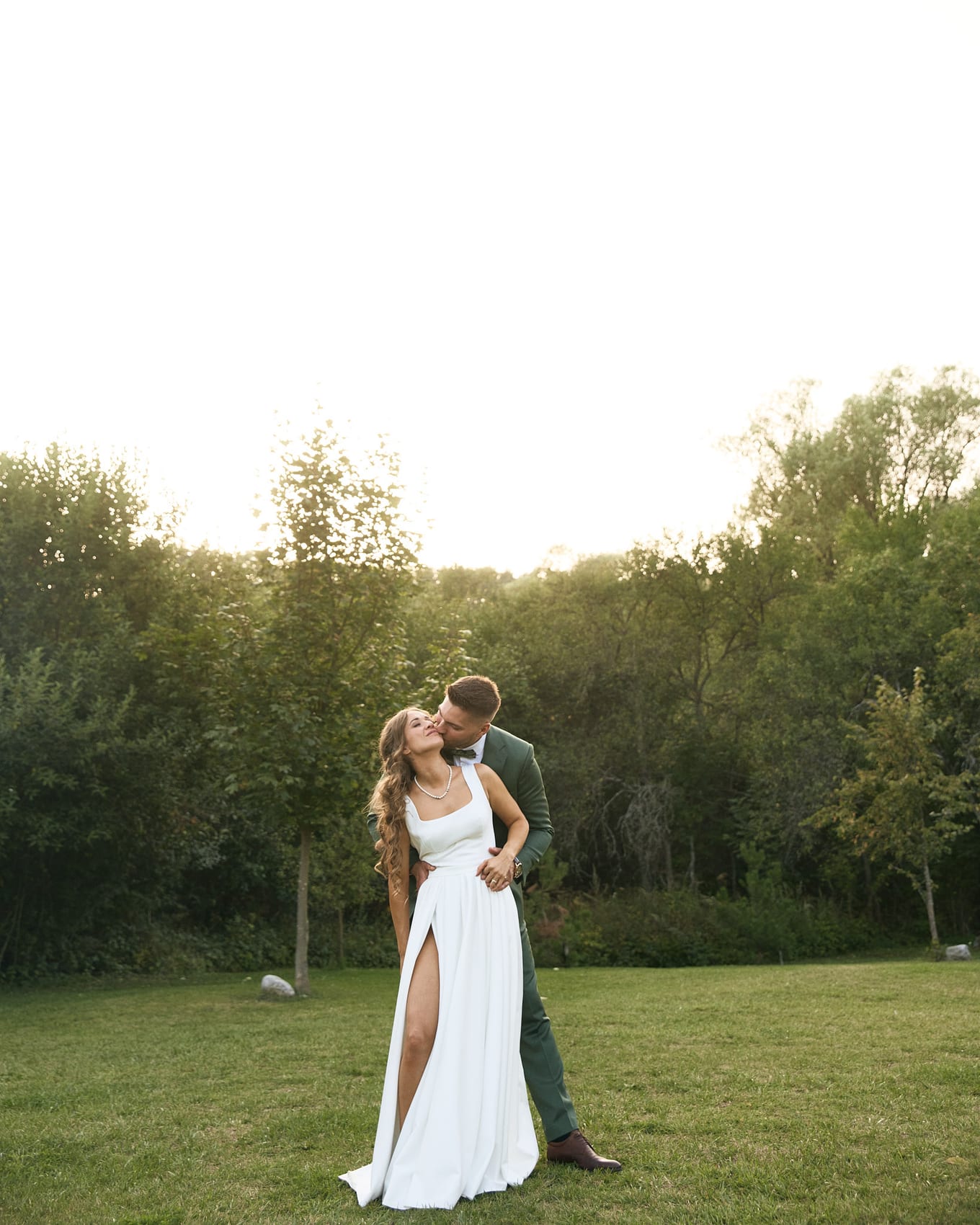 Groom kisses the bride's cheek from behind as they stand on a lawn below a pale sky and trees