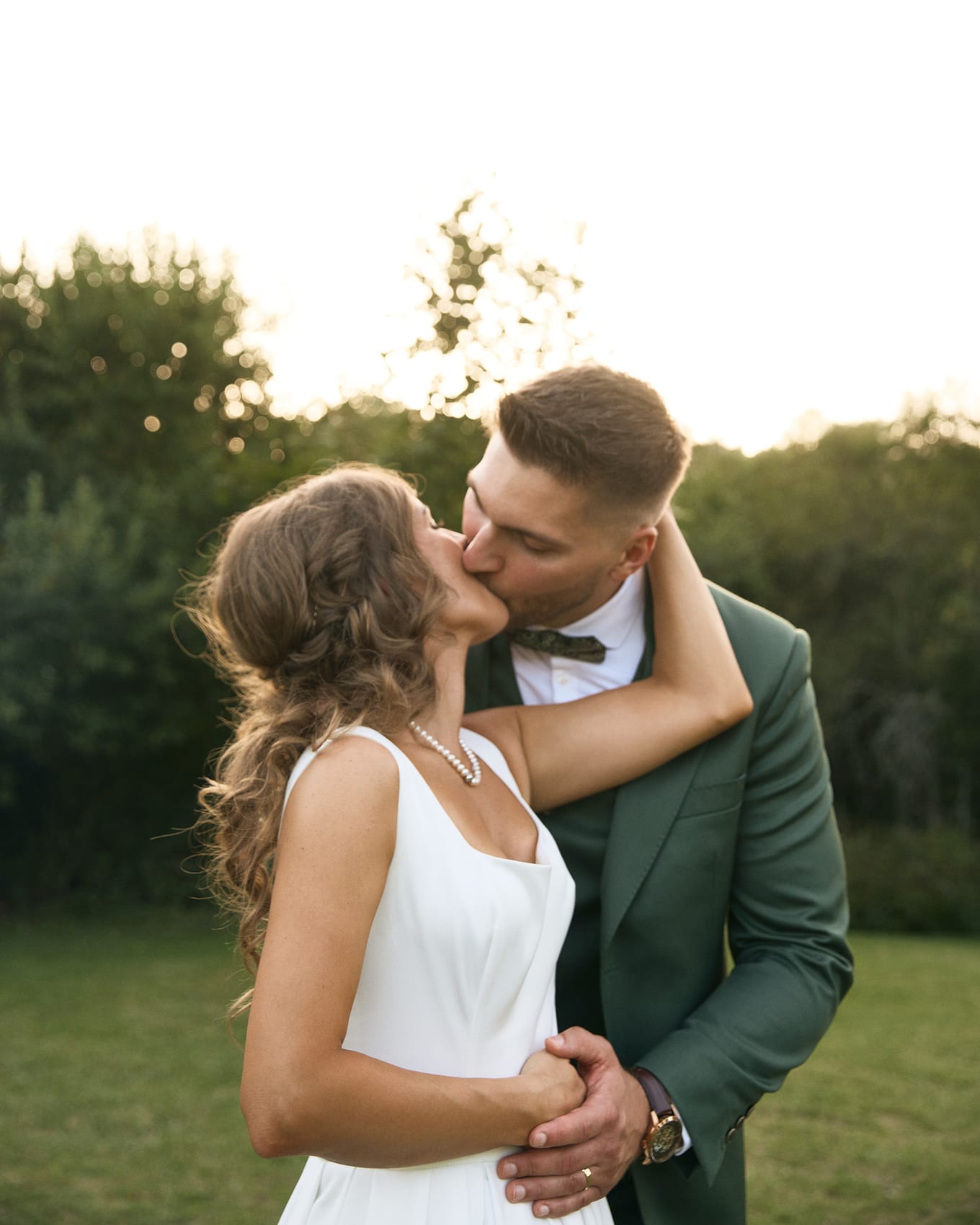 Kissing on a lawn at dusk, the bride's arm wrapped around the neck of a groom in a green suit