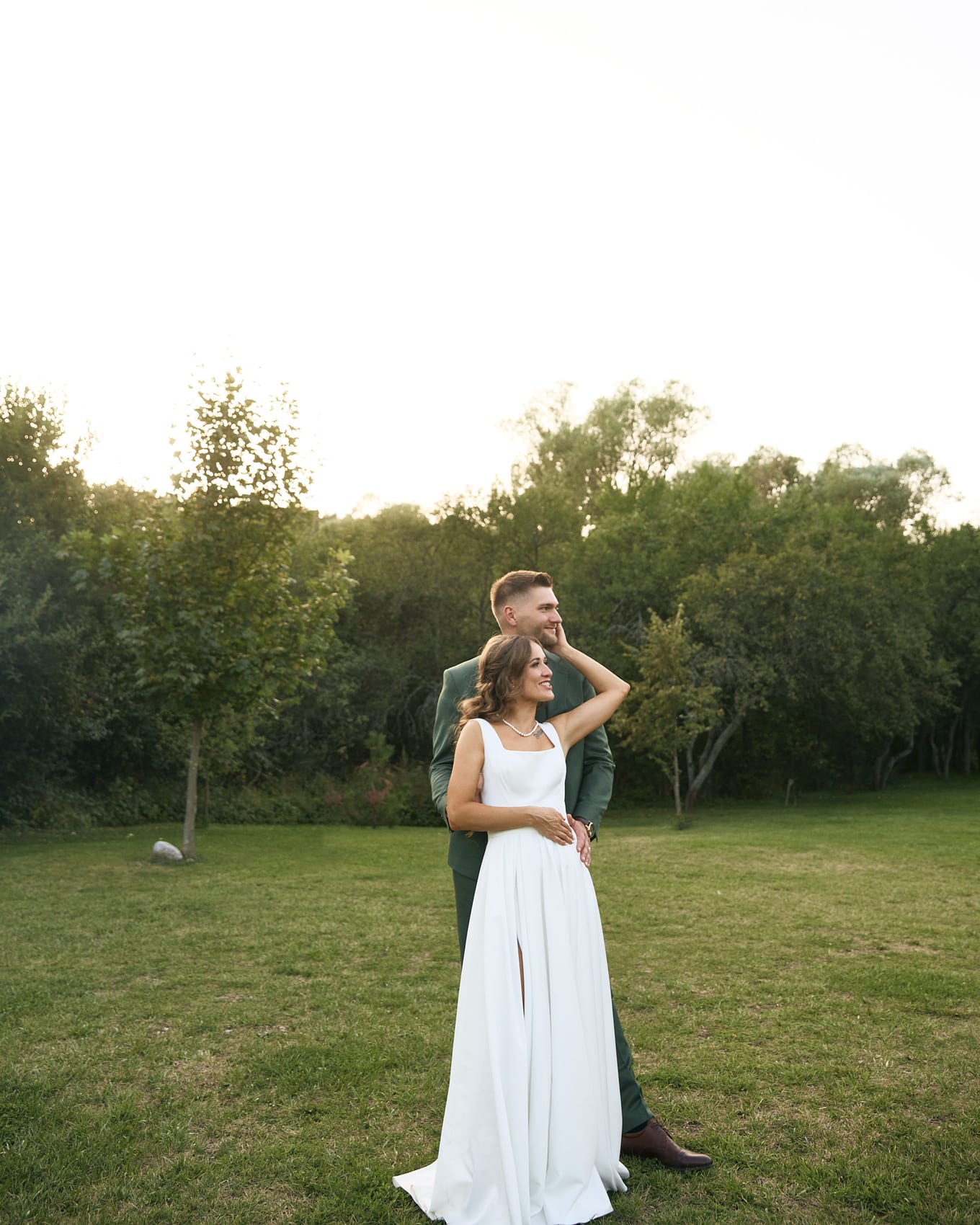 Groom in a green suit holds the bride from behind as they stand on a wide lawn edged with trees