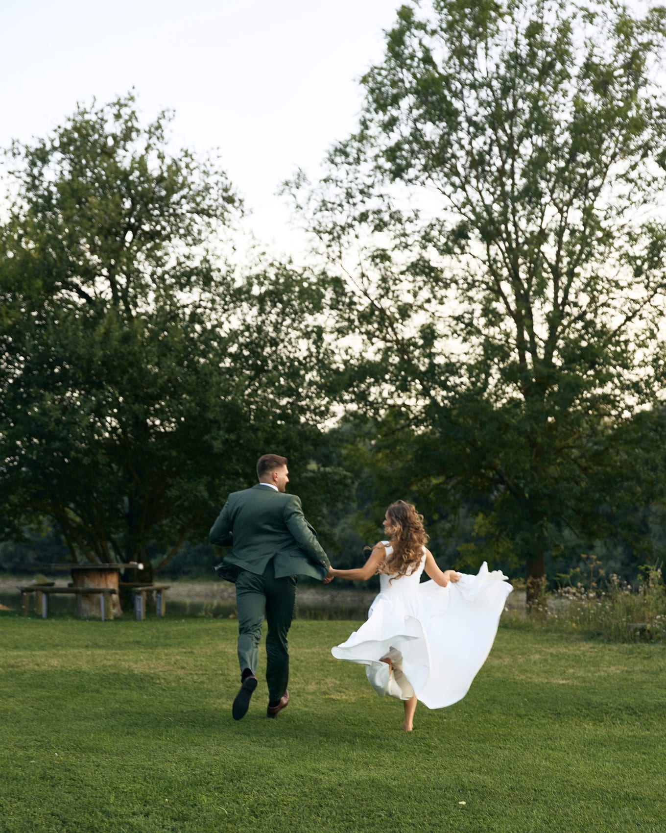 Seen from behind, a man in a green suit and a woman in a white gown dash over a lawn holding hands