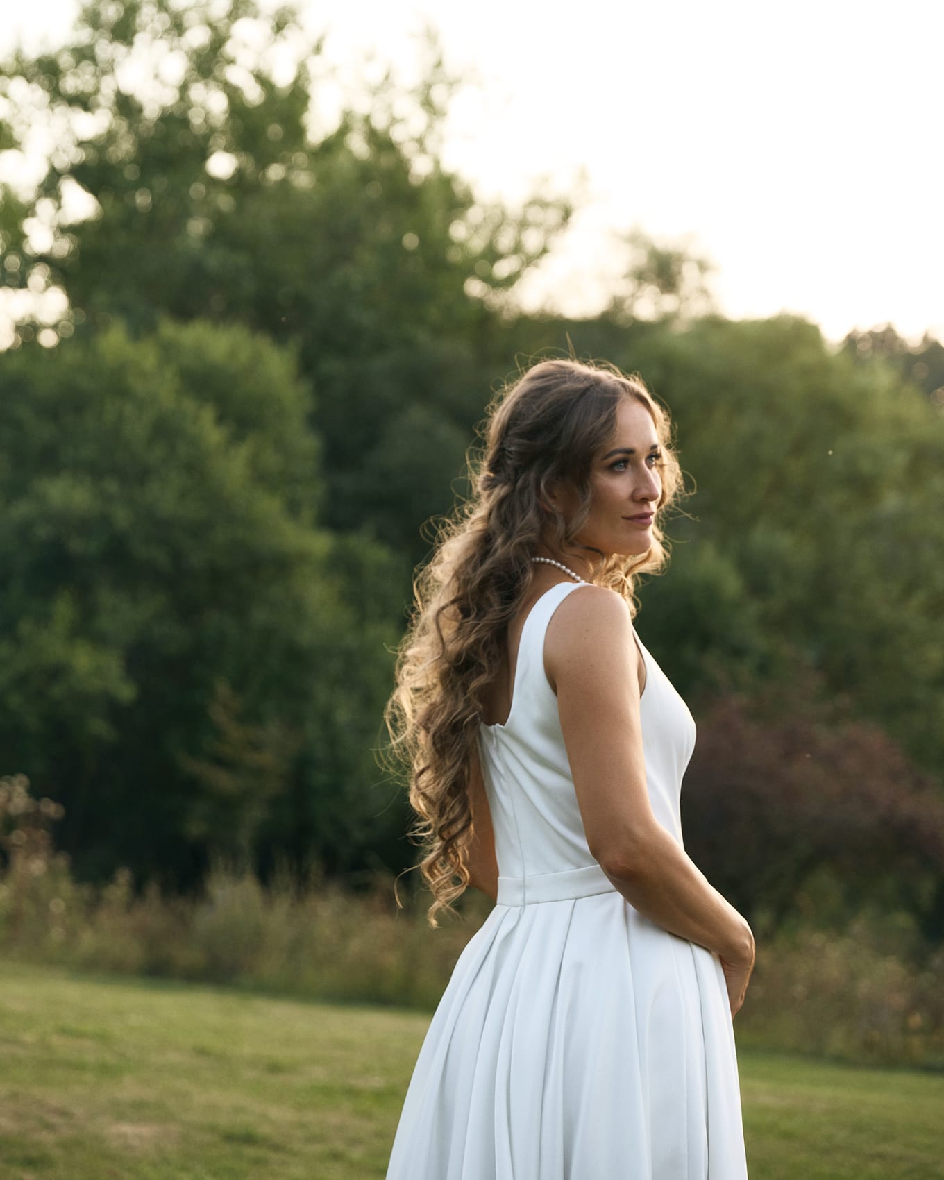 Long curly hair falls down the back of a woman in a sleeveless white dress glancing over her shoulder
