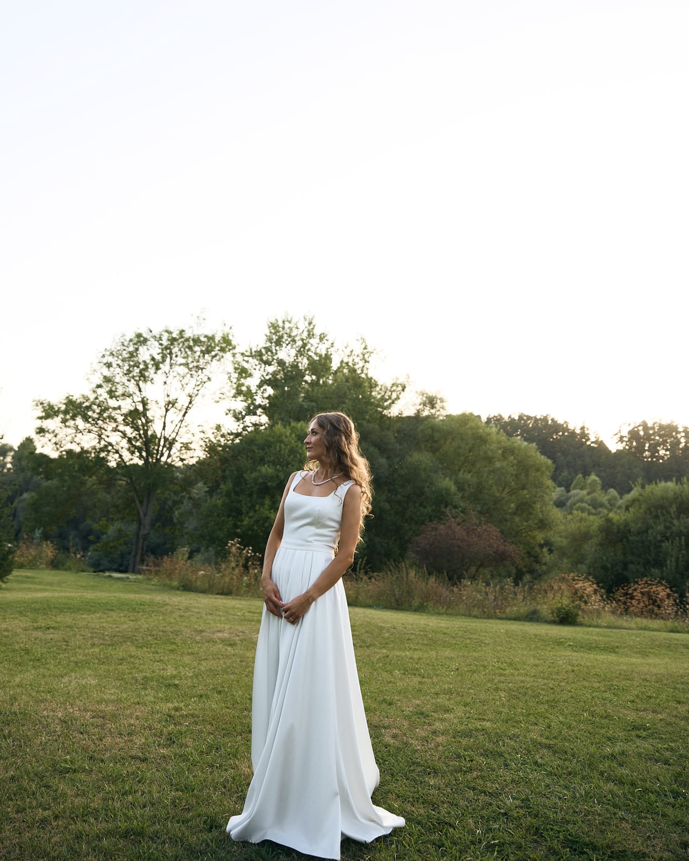 Bride in a sleeveless white dress and pearl necklace stands in a grassy field at dusk