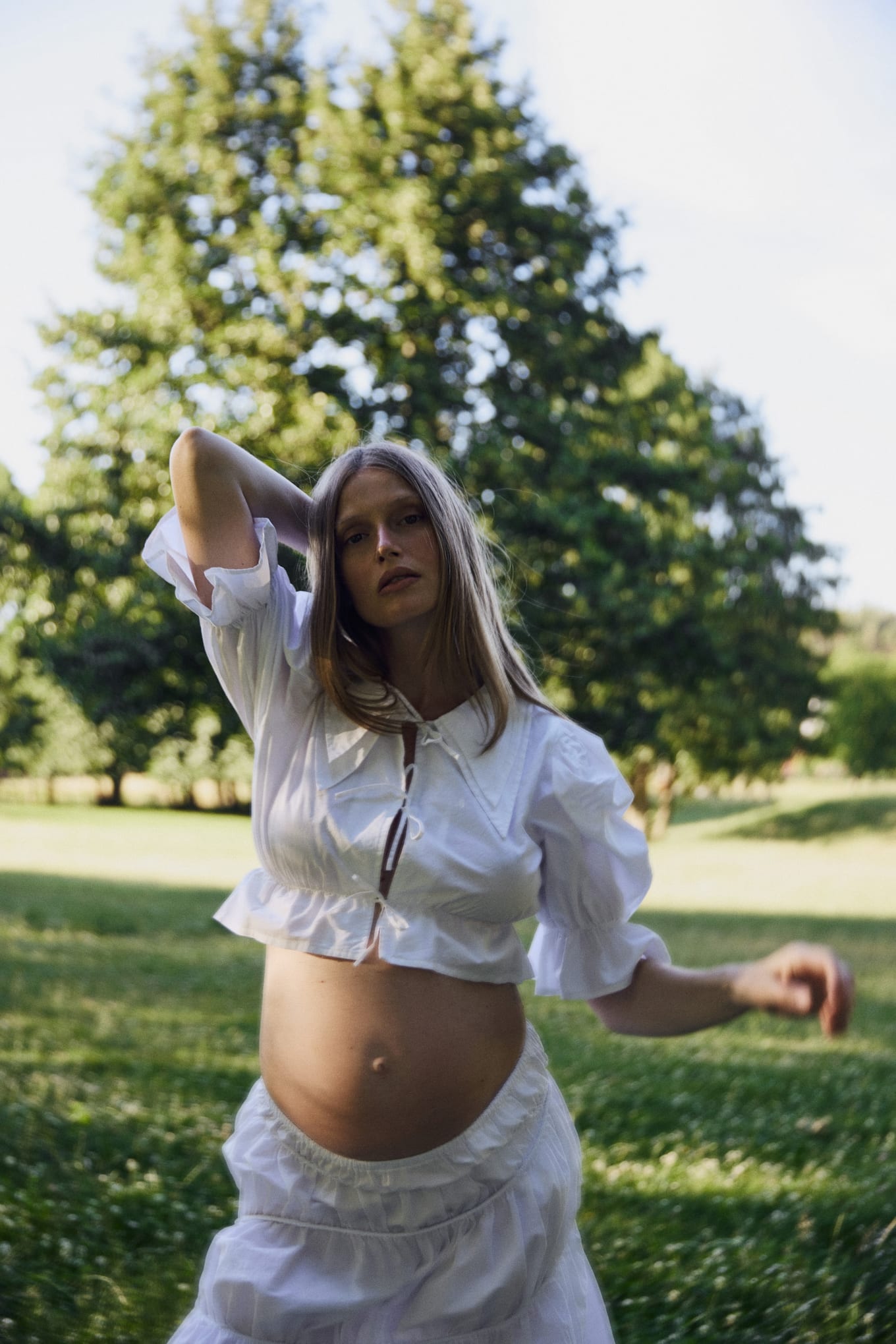 Woman with a bare pregnant belly raises one hand to her head beside a tall tree in a park