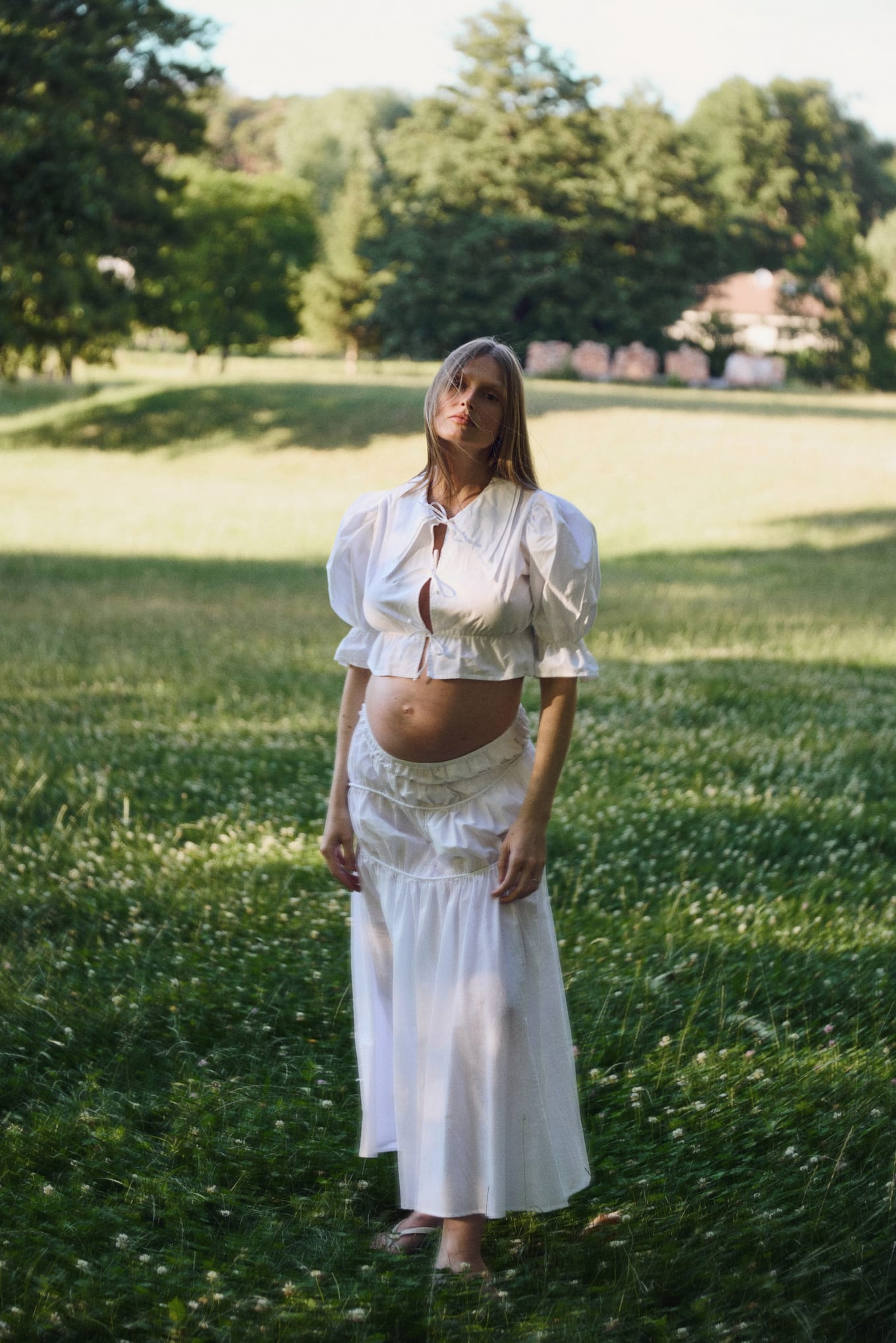 Standing barefoot in clover, a pregnant woman wears a white crop top and long tiered skirt