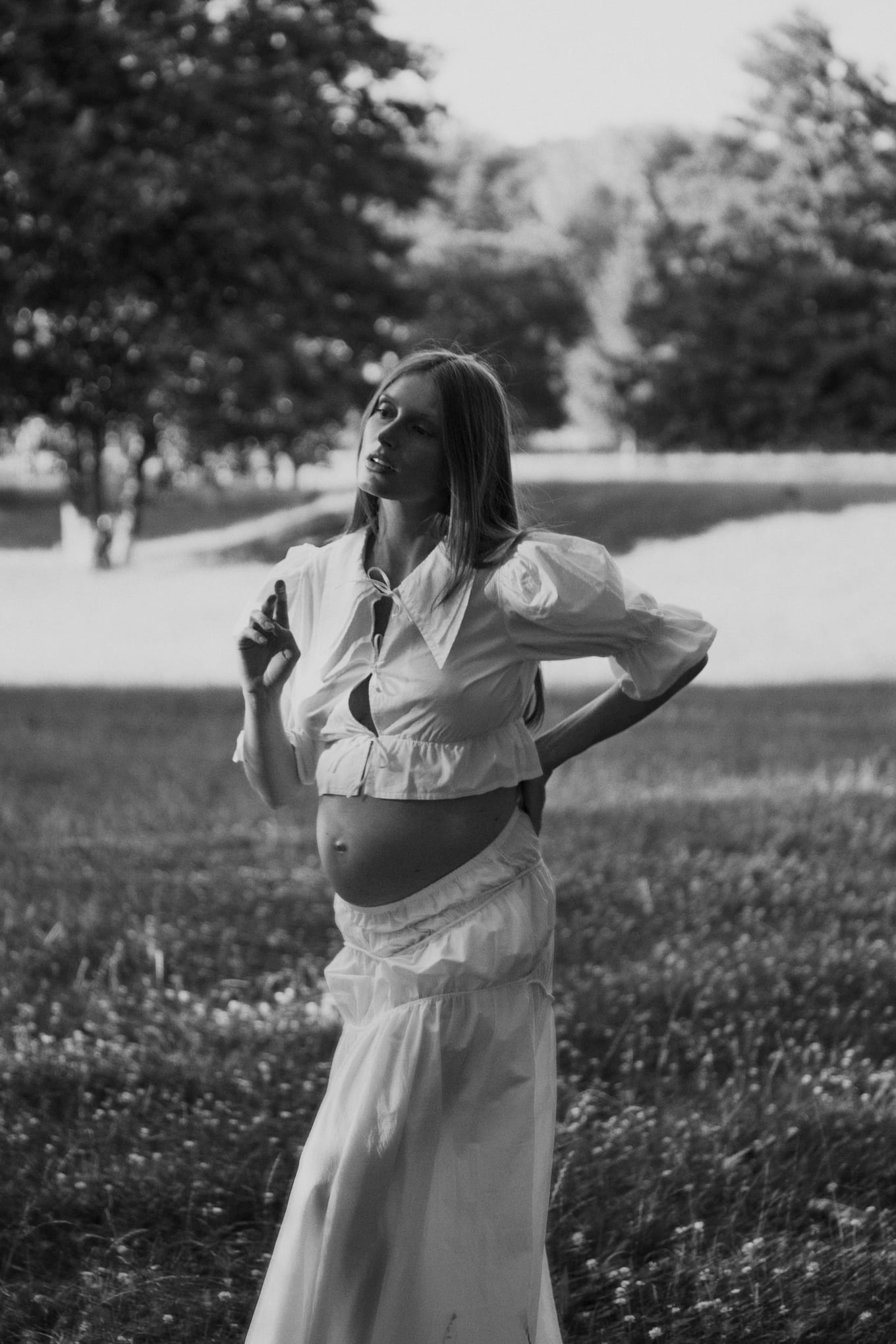 Black and white shot of a pregnant woman in white touching her collar in a grassy field