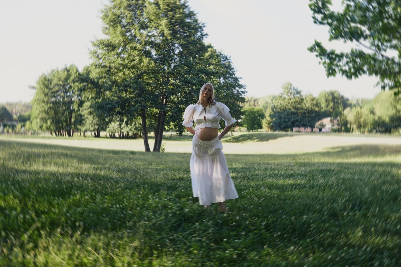 Pregnant woman in a white blouse and long skirt stands in a sunlit meadow with tall trees behind