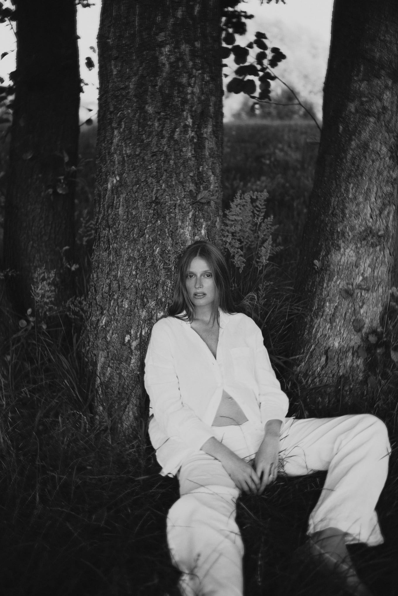 Woman in a white linen shirt and trousers sits against a tree trunk in tall grass, black and white