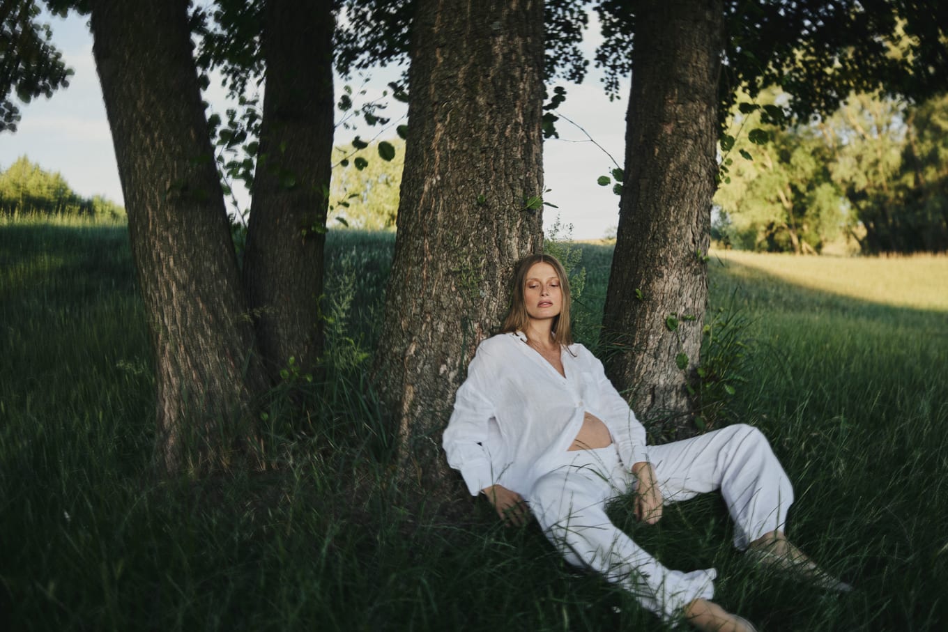Seated between tree trunks in evening sun, a woman in white rests with her eyes closed