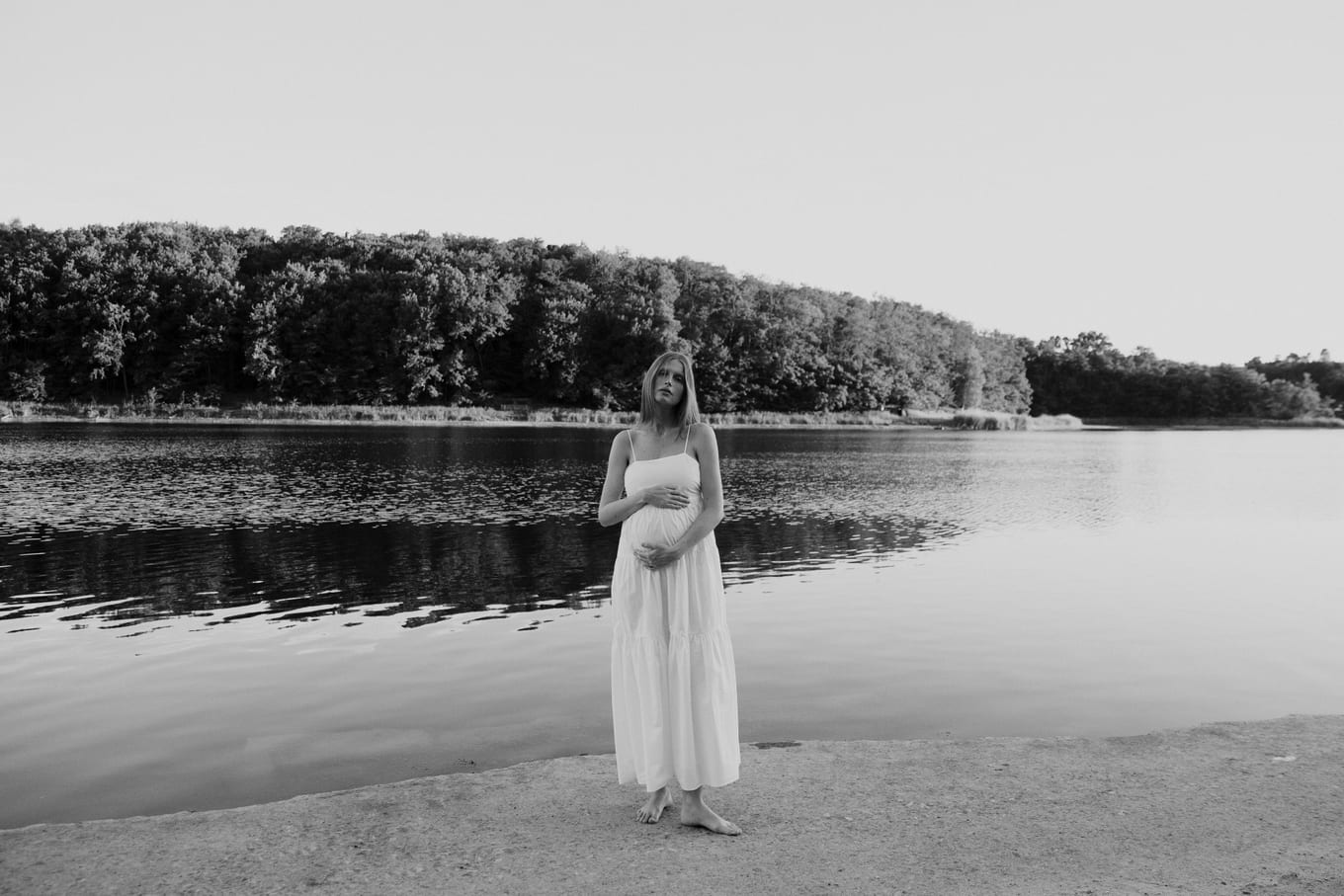 Barefoot on a lakeshore, a woman in a long white dress holds her belly with wooded hills behind