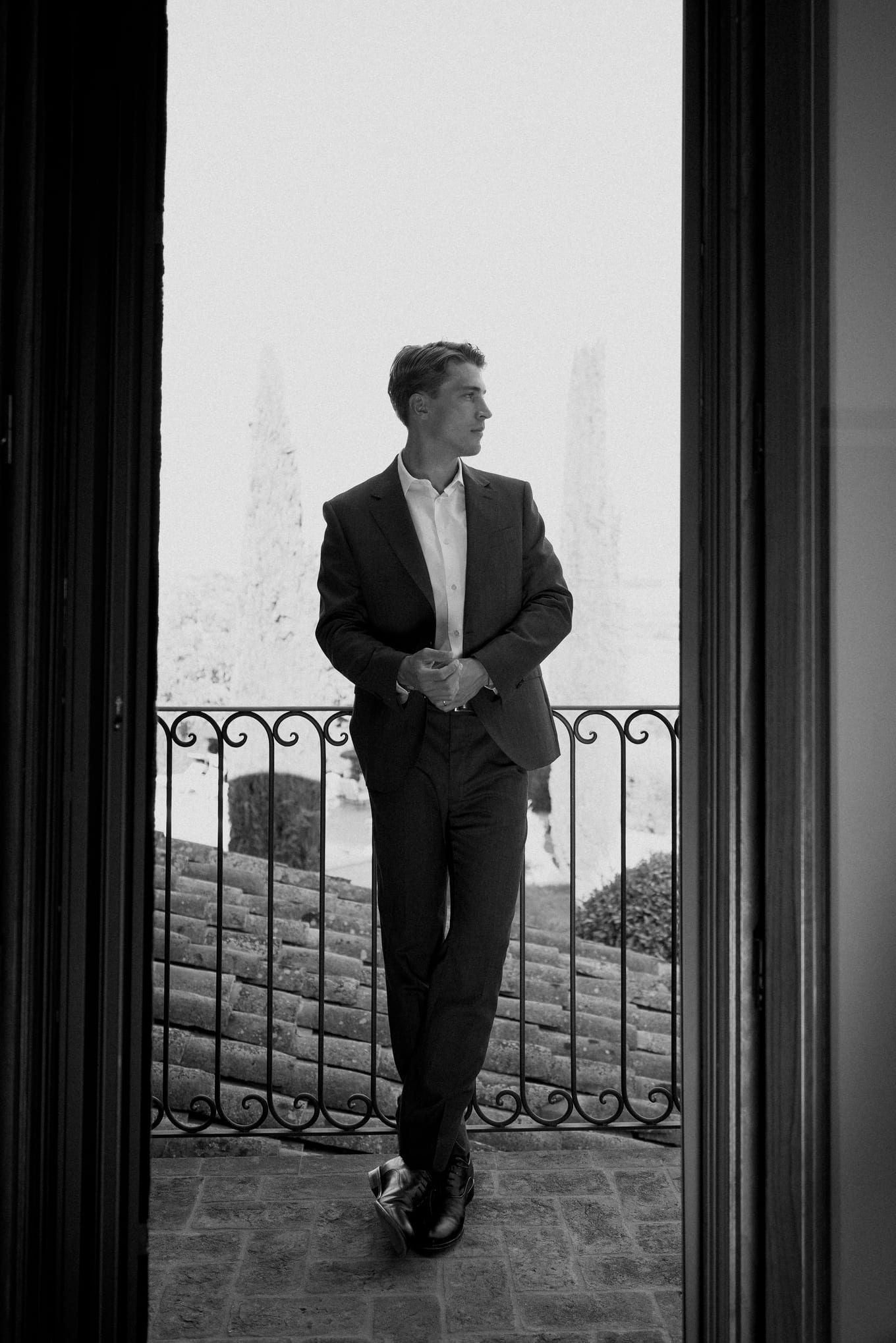 Framed by a doorway, a man in a dark suit leans against a wrought iron balcony railing above tiled roofs