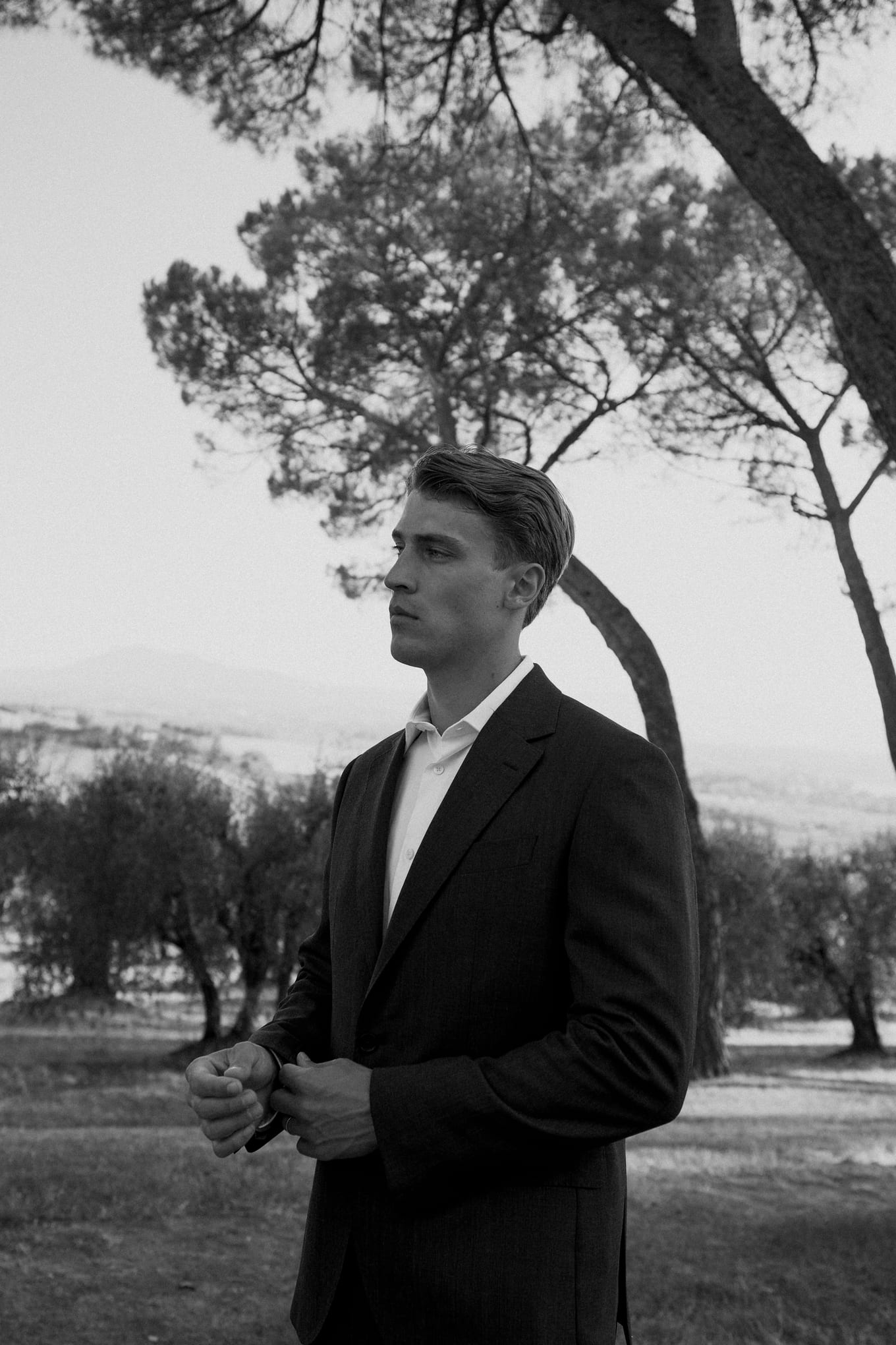Young man in a dark suit stands under umbrella pines, hands clasped, looking off to the side