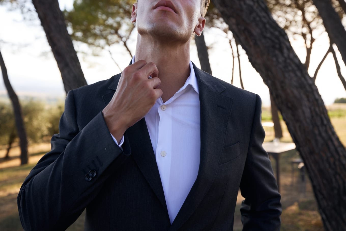 Cropped view of a man in a pinstripe suit adjusting his open white shirt collar beneath tall pines