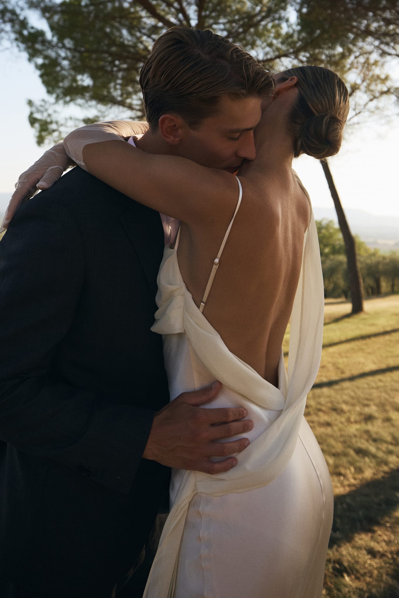 Man in a dark suit kisses the neck of a woman in a backless white satin dress under a pine tree