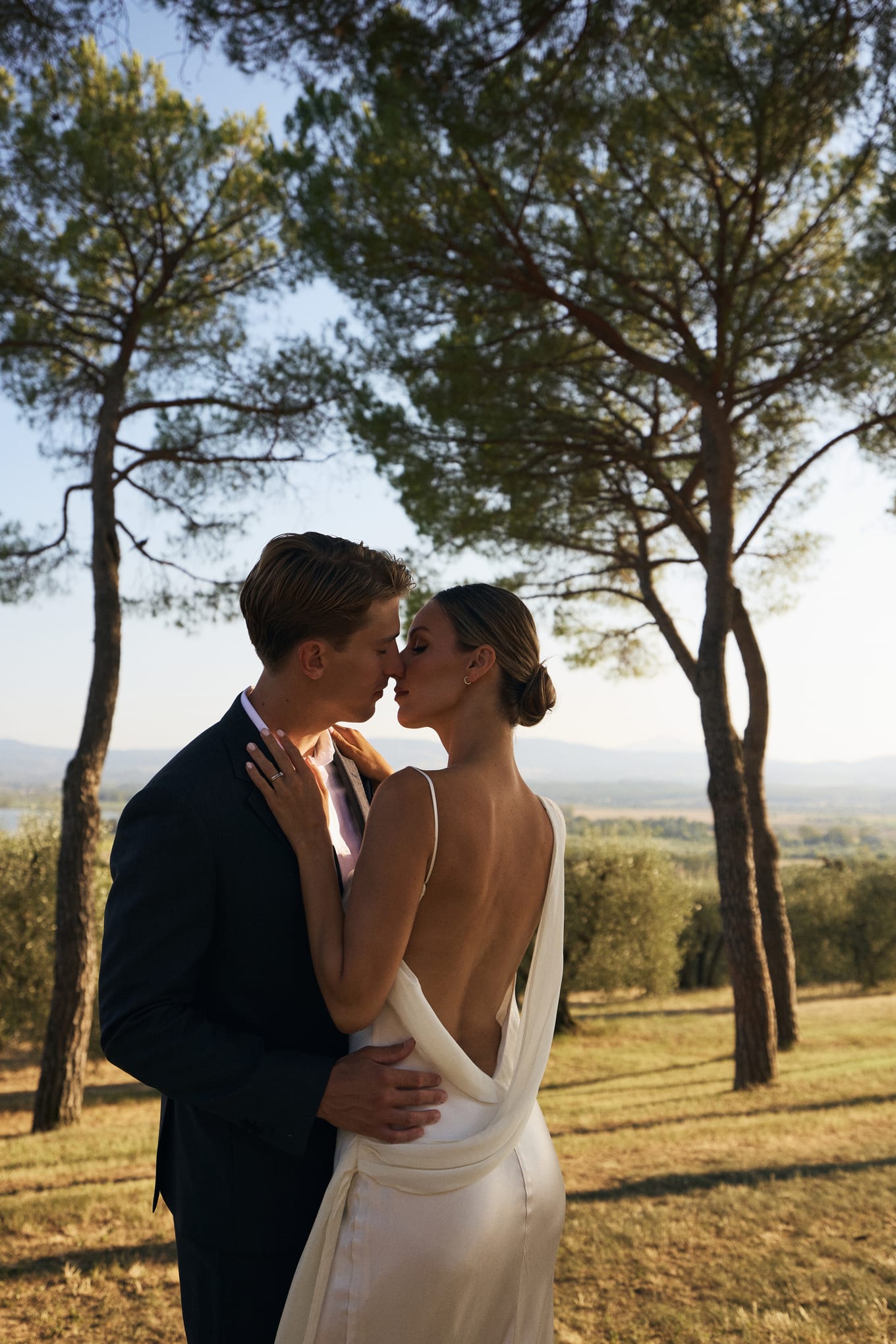 Under tall pines, a couple kiss as she rests a ringed hand on his shoulder, hills behind them
