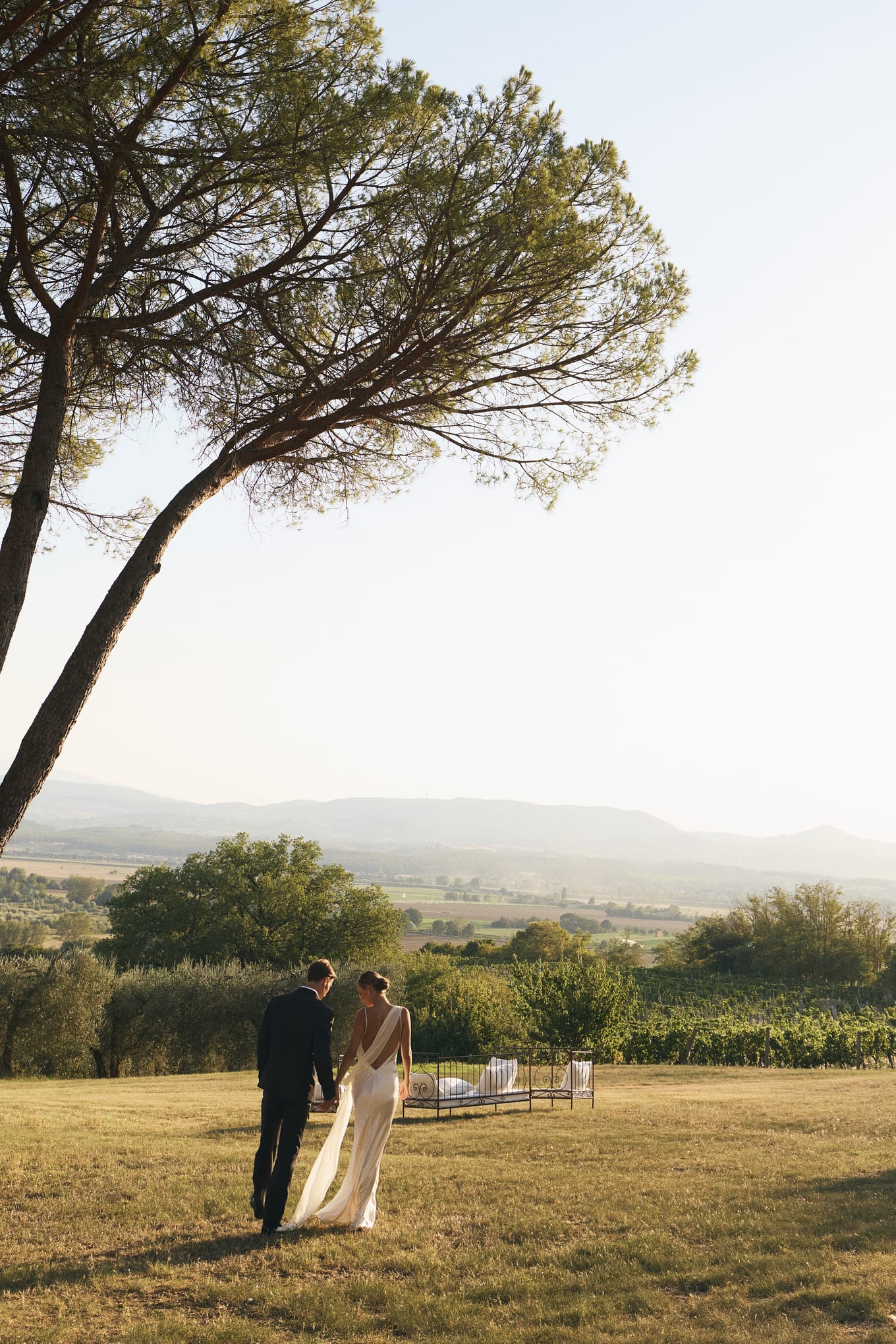 Holding hands, a man and a woman in a white gown walk across a sunlit lawn toward vineyards and hills