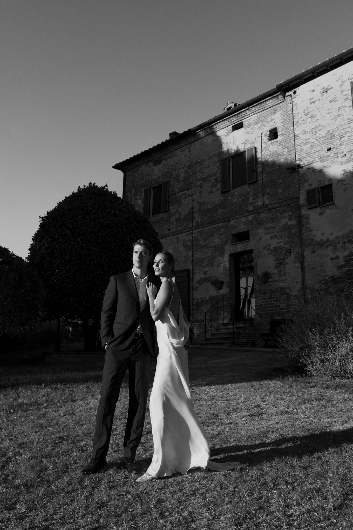 Man in a suit and woman in a white gown standing on grass before a tall brick villa, in black and white