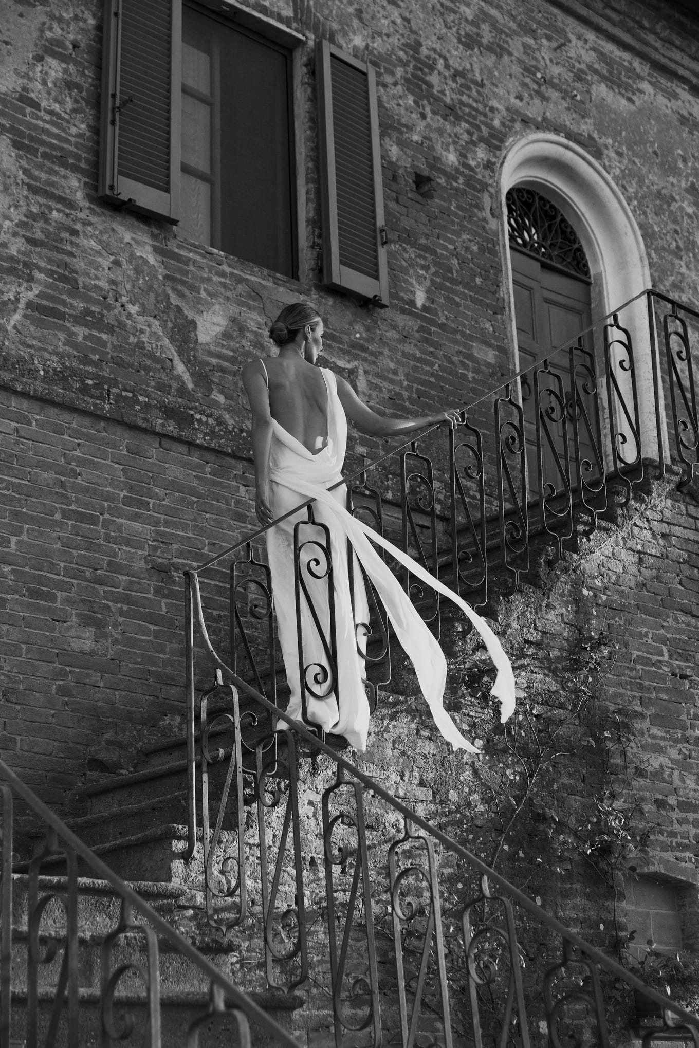 Bride in a backless gown climbing iron-railed stone stairs as her long sash blows out, in black and white