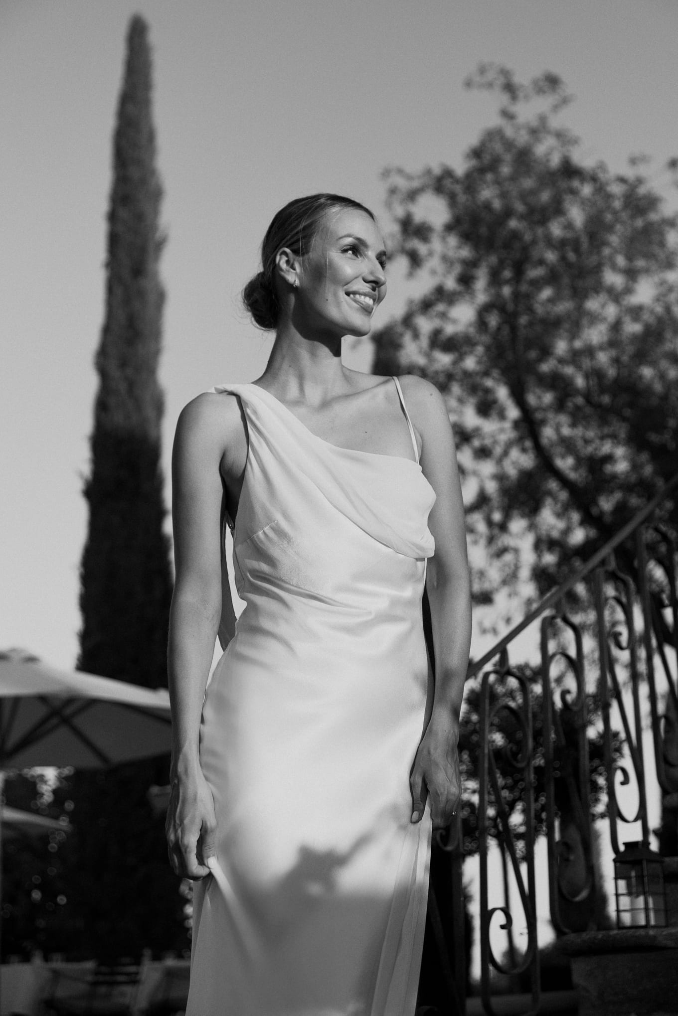 Smiling bride in a satin one-shoulder gown stands beside a wrought-iron railing with a cypress behind her
