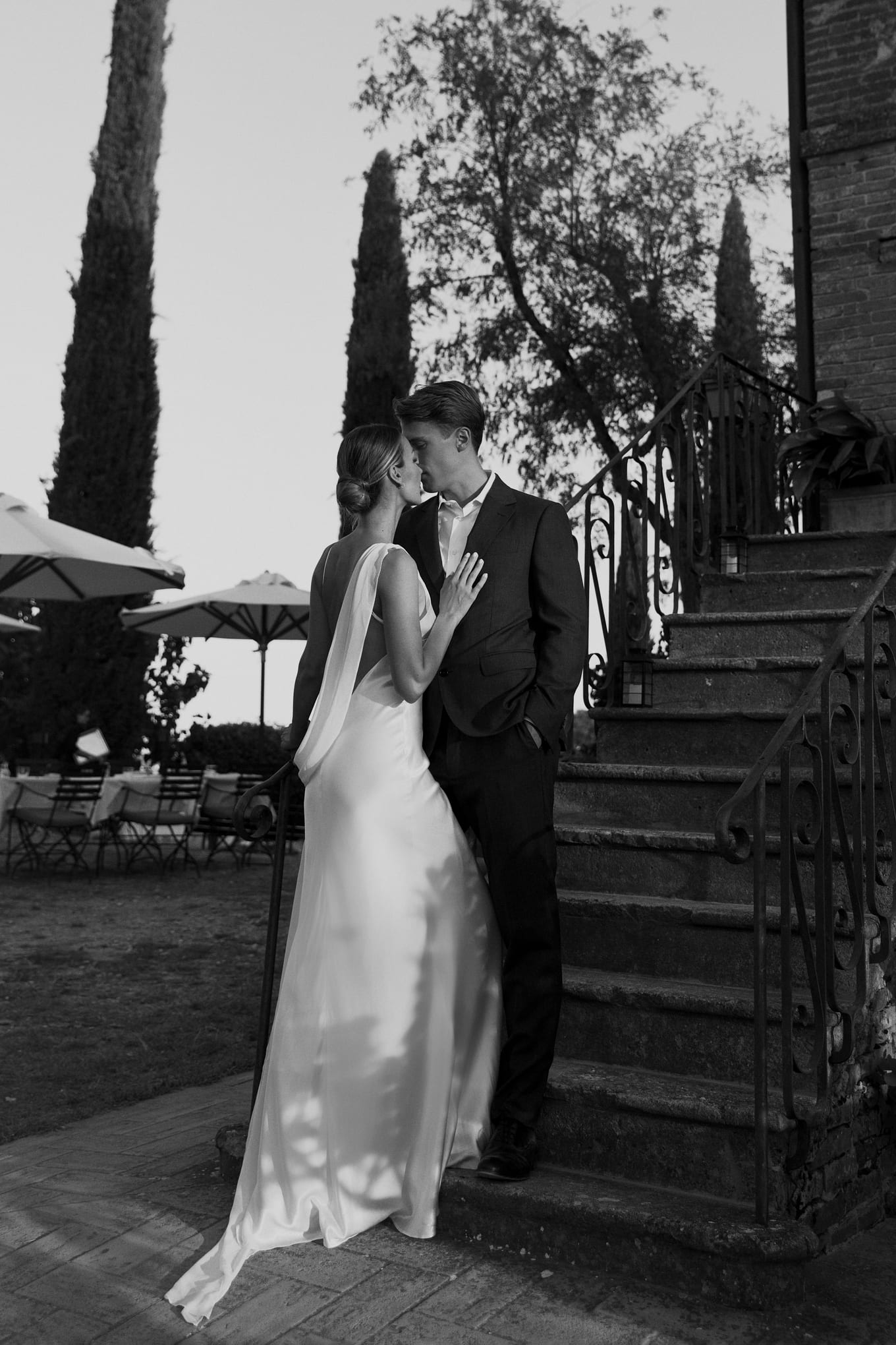 In black and white, a bride and groom kiss at the foot of stone steps with an iron railing