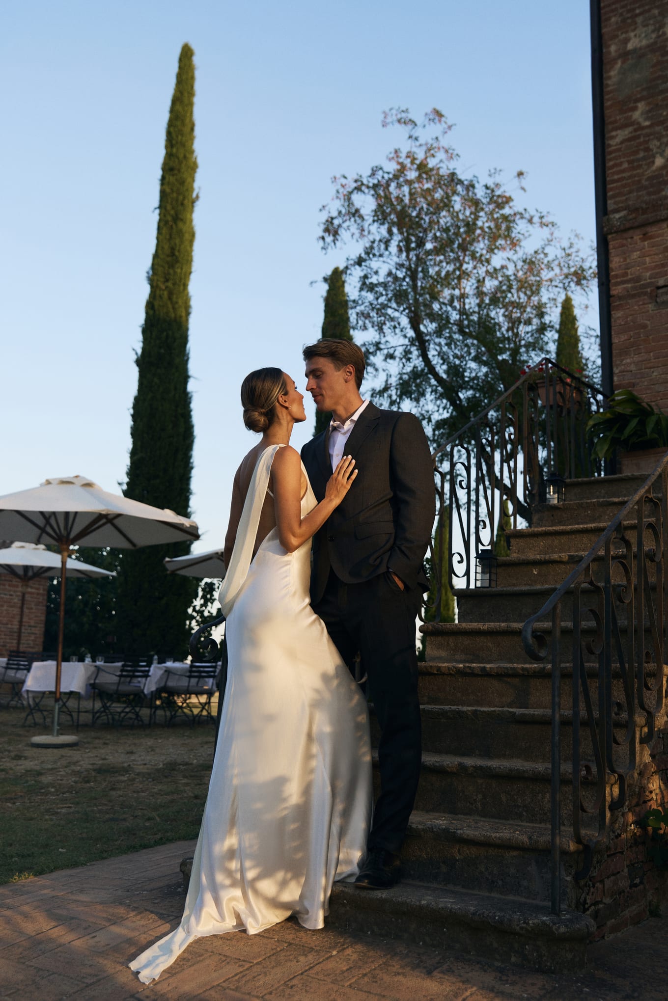 Bride in a satin gown and groom in a dark suit kiss by stone steps at golden hour, cypress trees behind