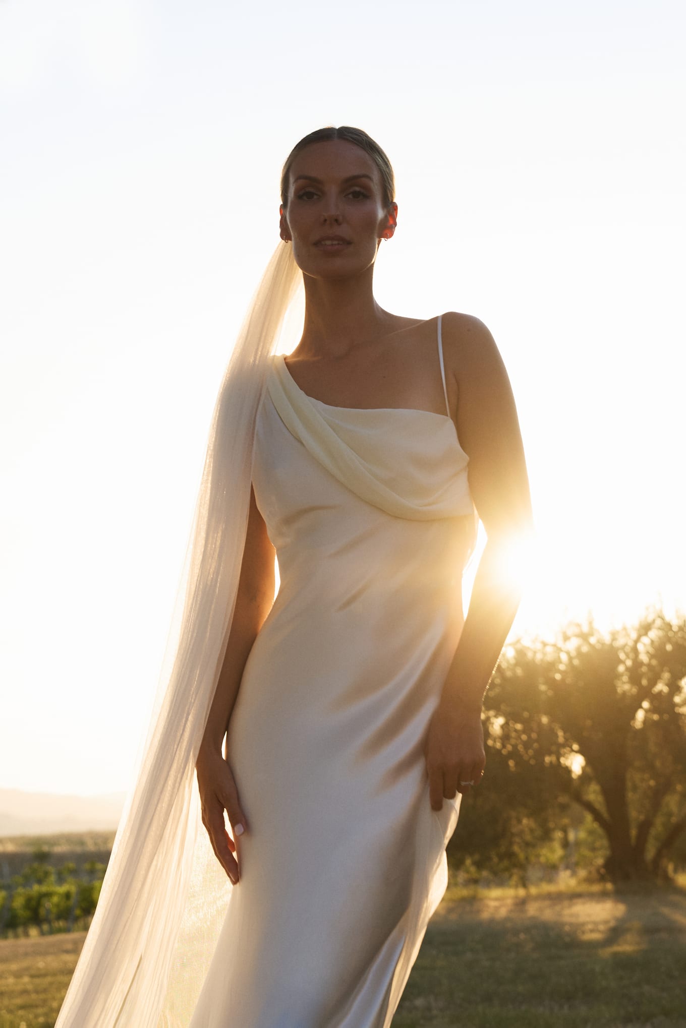 Close portrait of a bride in a cowl-neck satin dress and veil, facing the camera with the sun behind her