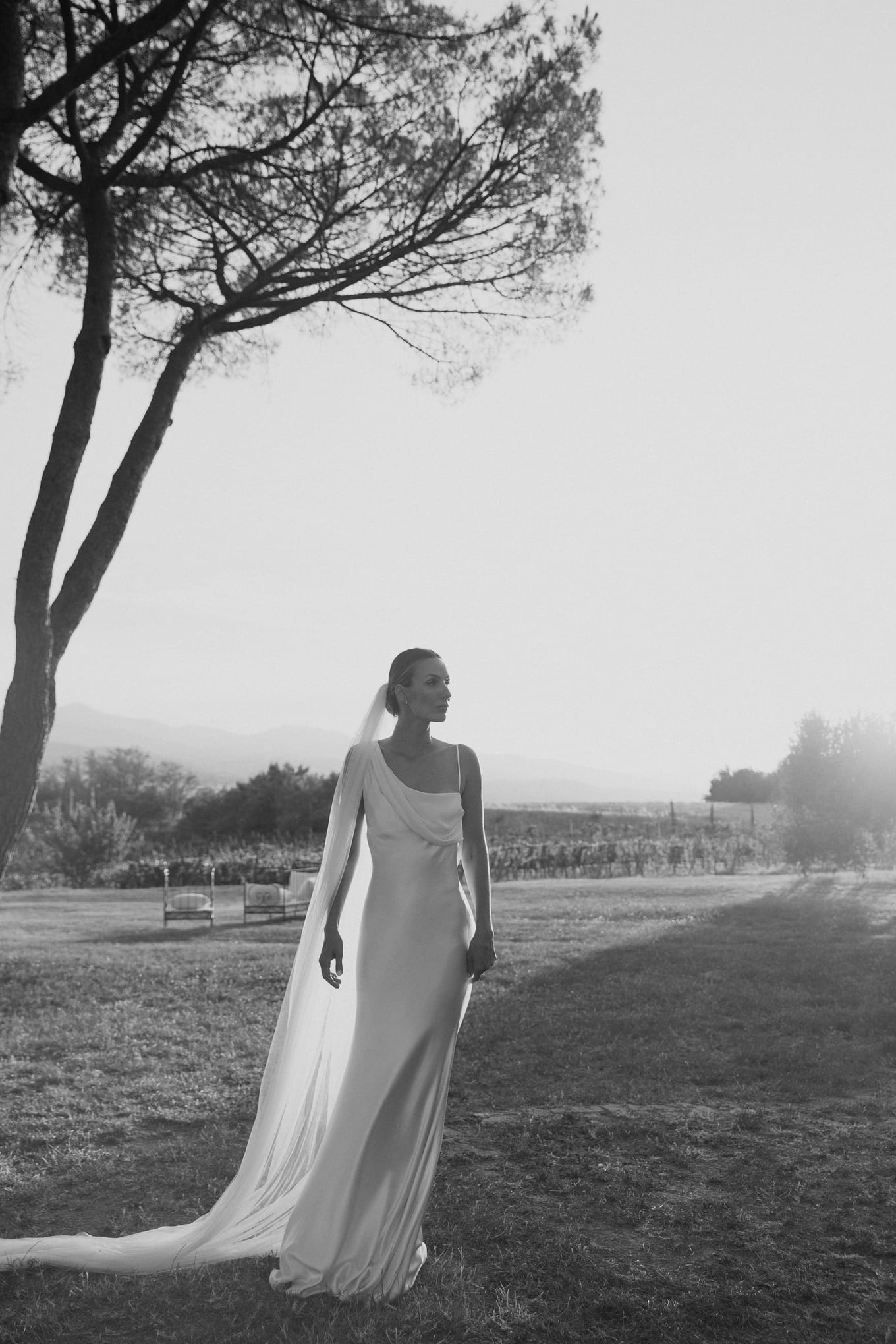 Black and white view of a bride in a draped satin gown and long veil standing on a lawn beneath a pine