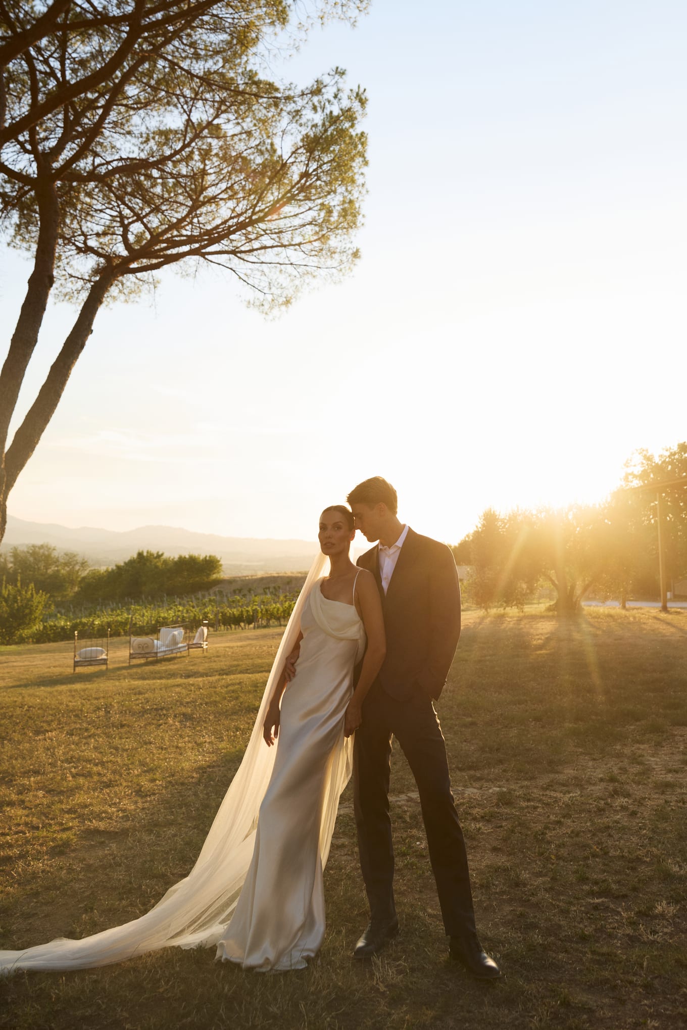 Groom in a dark suit leans his head against a bride in a satin gown on a sunlit lawn with vineyards behind