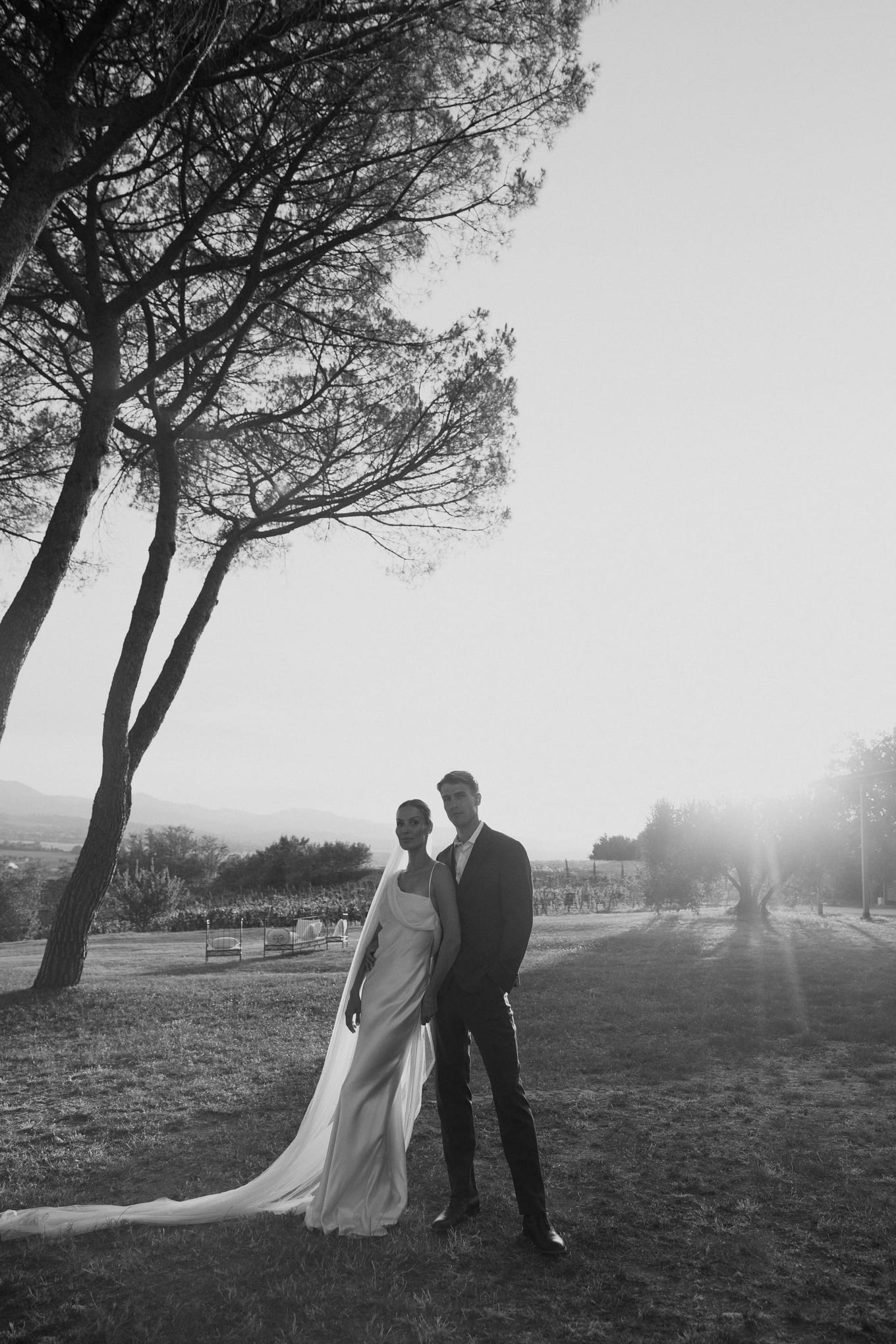 Bride in a long veil and groom stand together under tall pines on a lawn in black and white