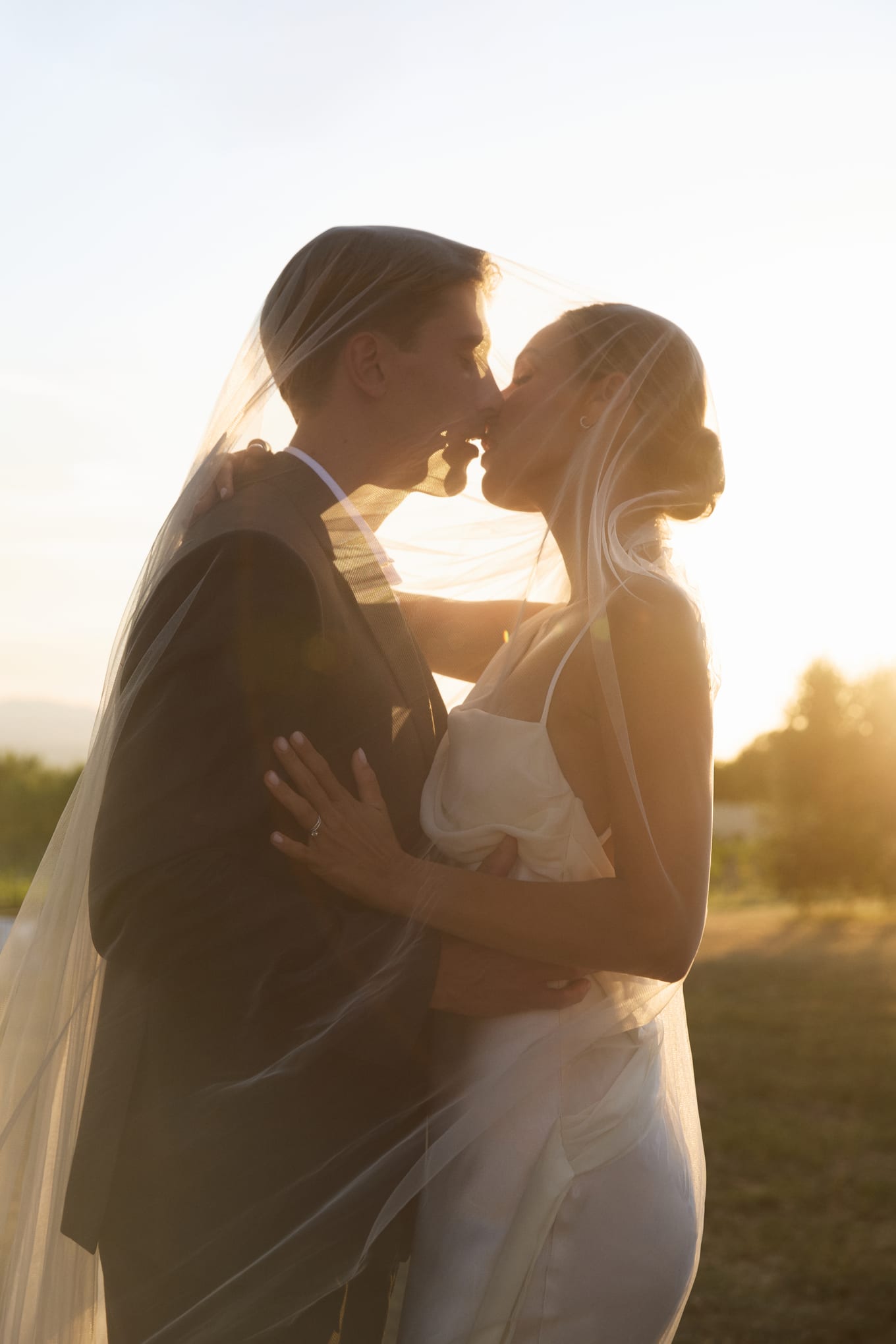 Under a sheer veil, a man in a dark suit kisses a woman in a white dress against the setting sun