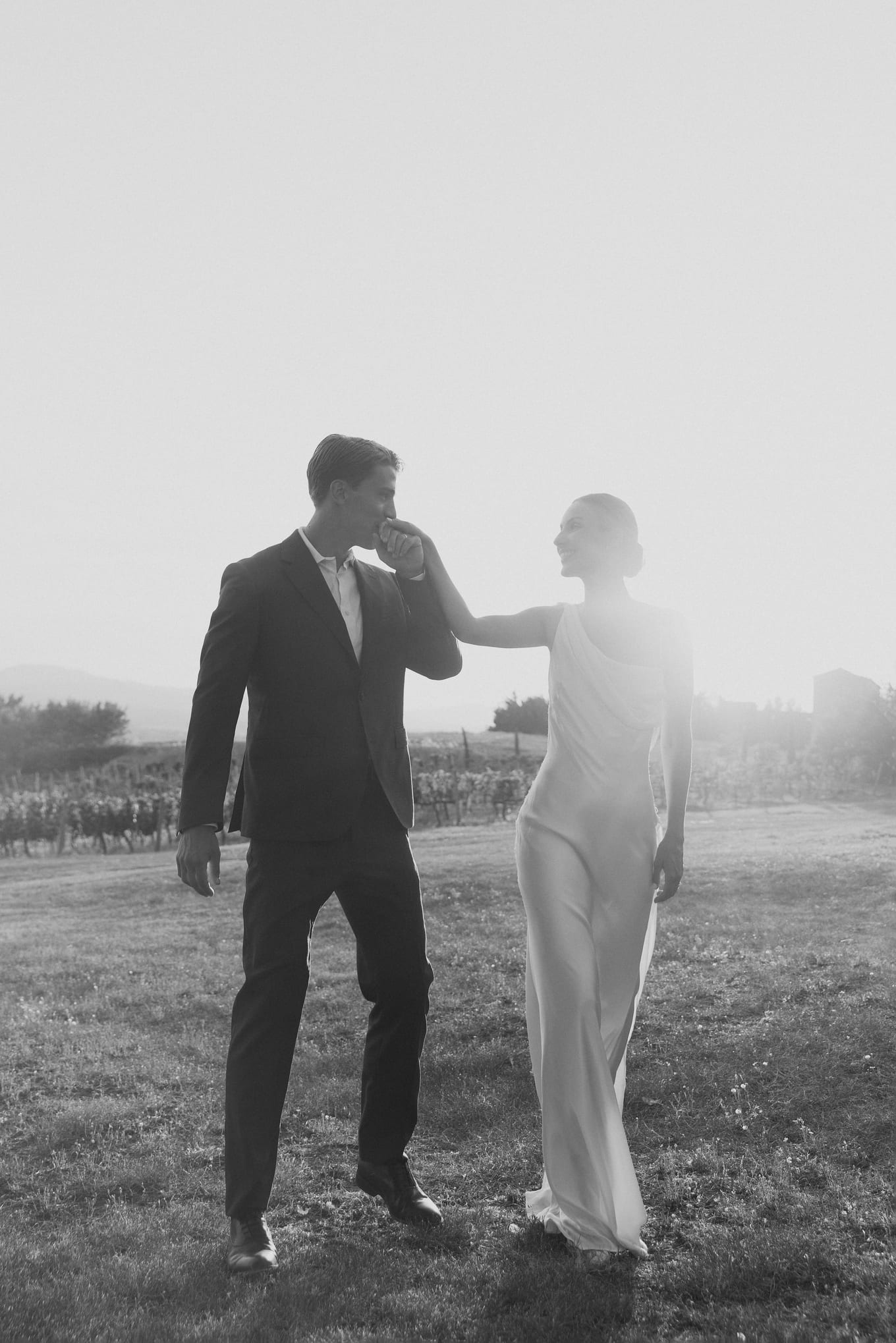 Groom in a dark suit kisses the bride's hand as they walk across a sunlit field in black and white