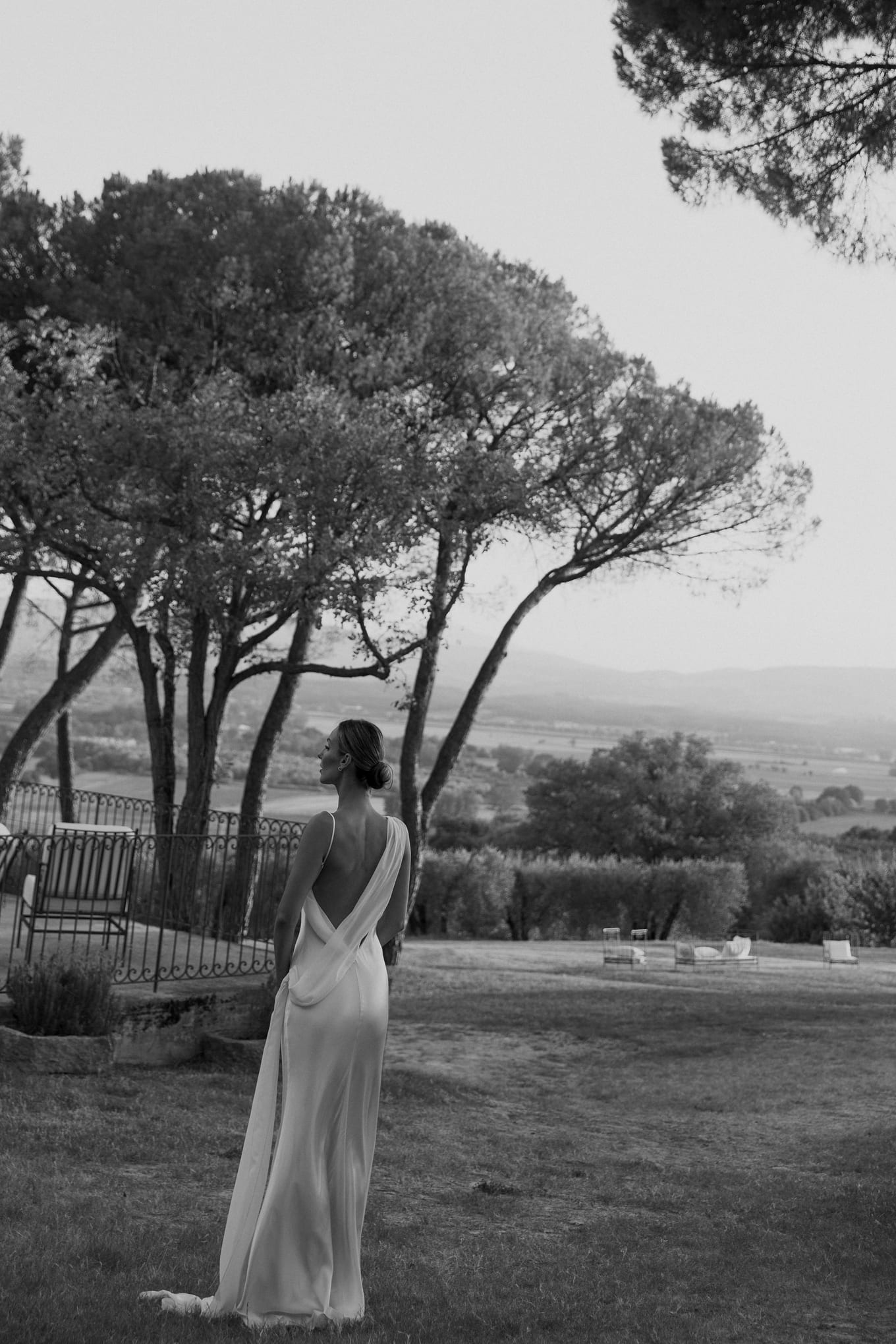 In black and white, a bride stands on a lawn beneath tall pines, her open-back dress trailing behind