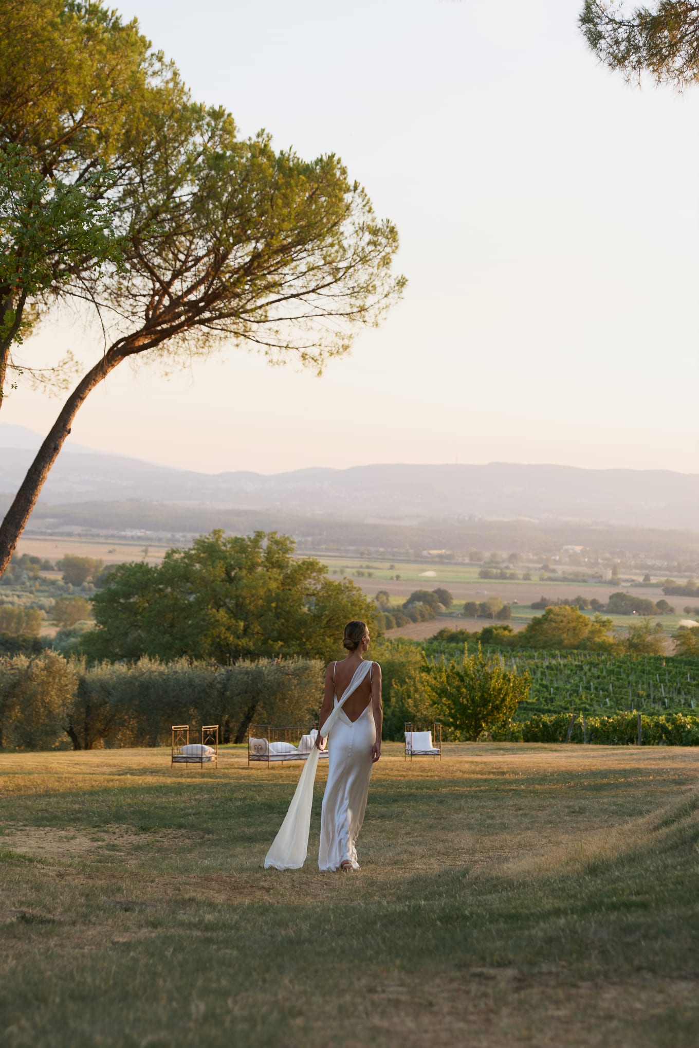 Seen from behind, a bride walks across a grassy lawn toward lounge chairs and vineyards at sunset