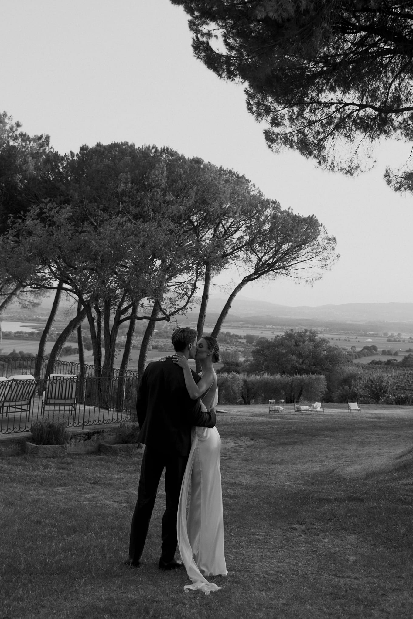 Under tall pine trees, a man in a dark suit kisses a woman in a long white gown on the grass