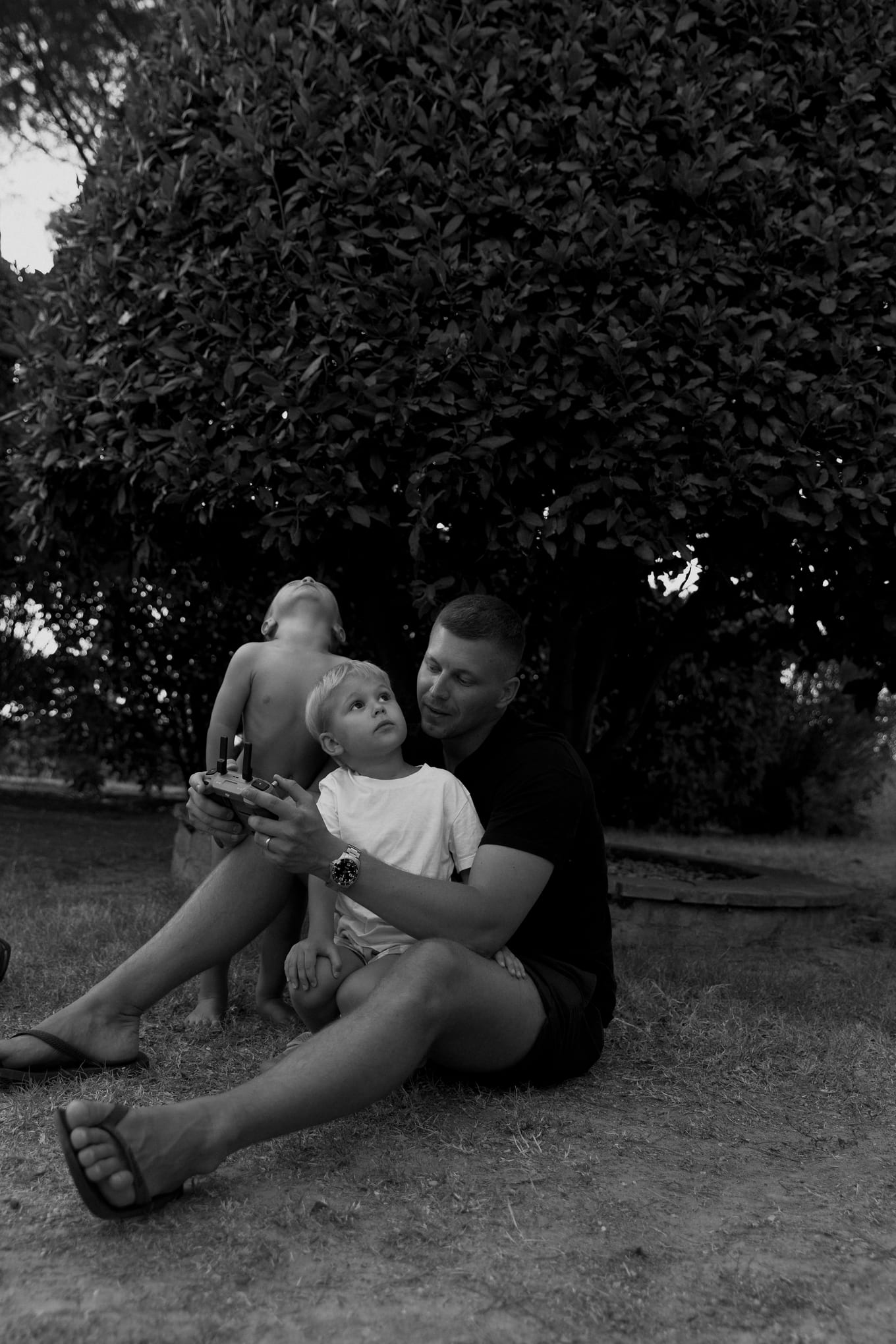Seated on the grass under a large shrub, a man holds a small boy while another child looks up